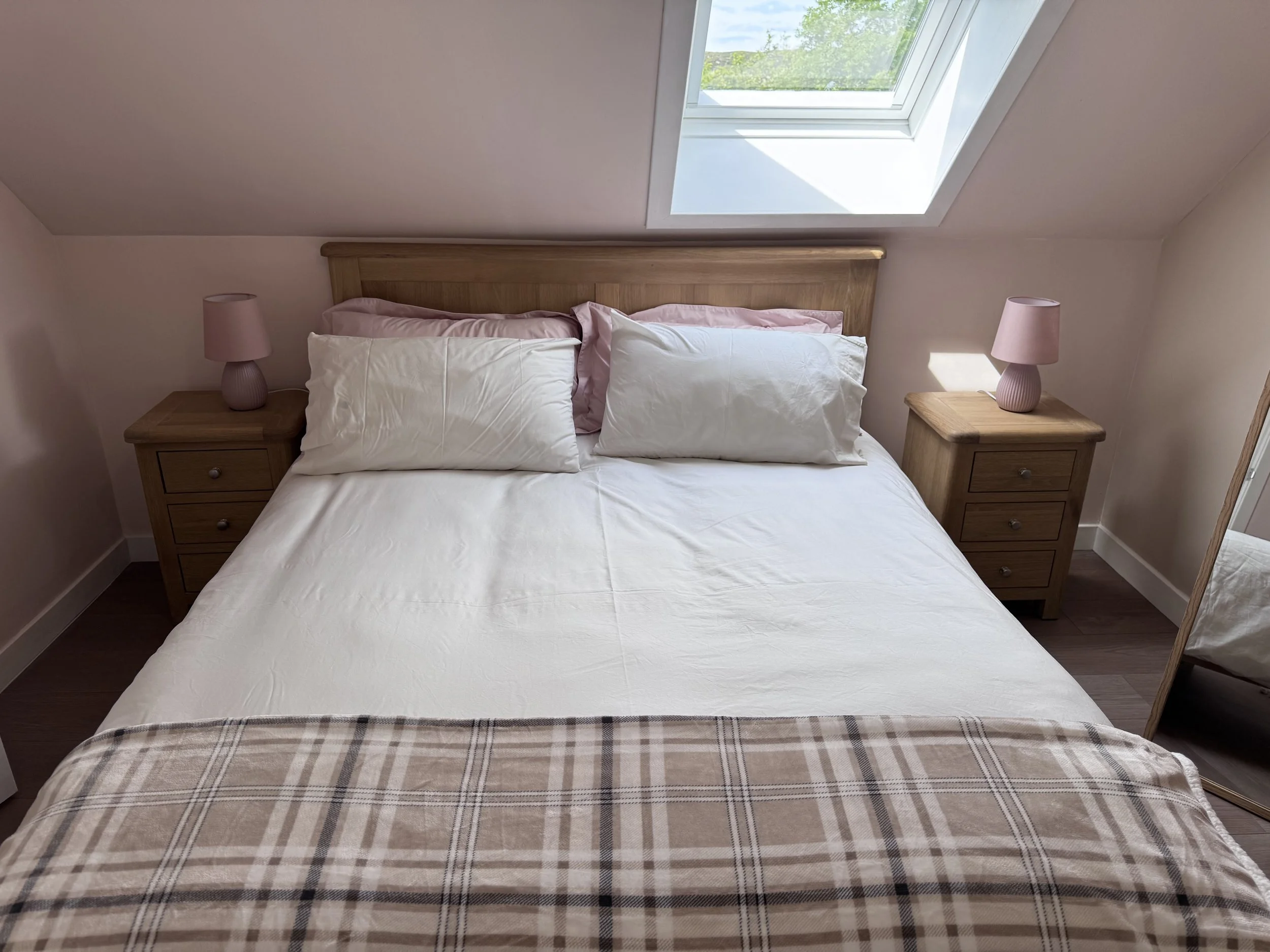A bedroom with a wooden bed frame, white bedding, pink and white pillows, two wooden nightstands with pink lamps, and a large skylight window showing green trees outside.