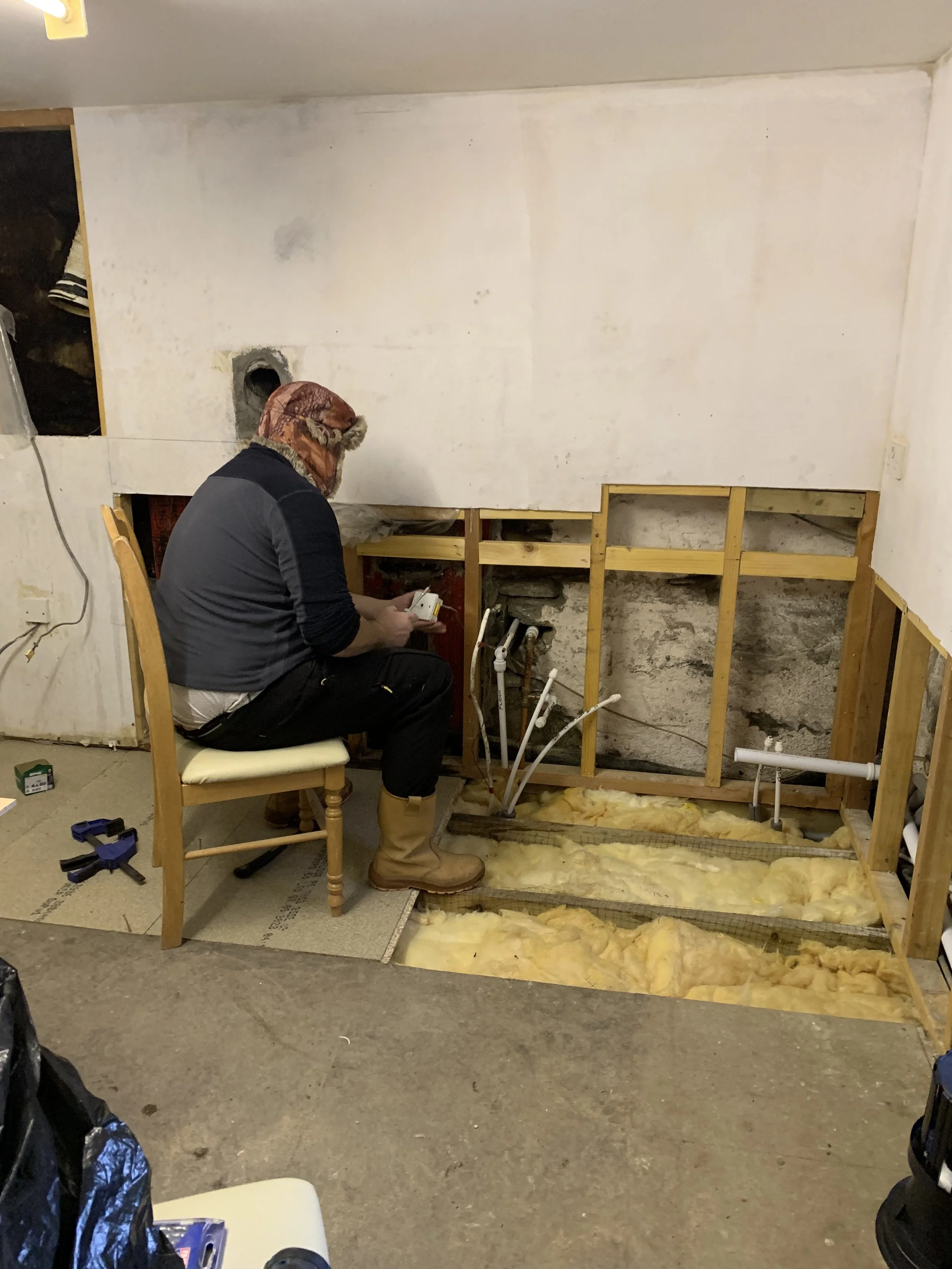 A person wearing a patterned hoodie, black jacket, black pants, and tan boots kneeling and working on plumbing behind a partially removed wall with exposed pipes and insulation. The room appears to be under renovation.