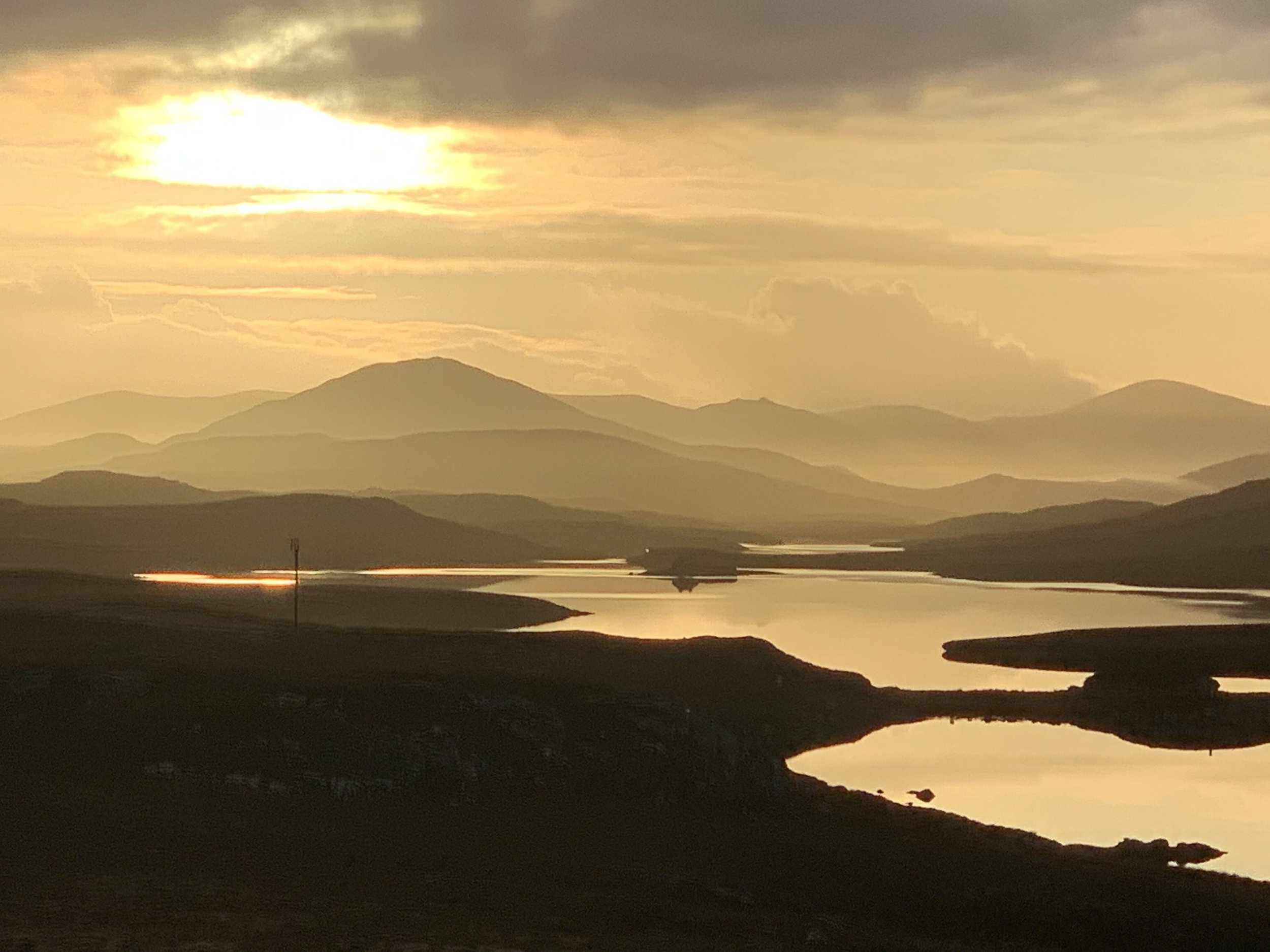 A landscape of mountain ranges during sunset with reflective bodies of water in the foreground and a cloudy sky with the sun partially visible.