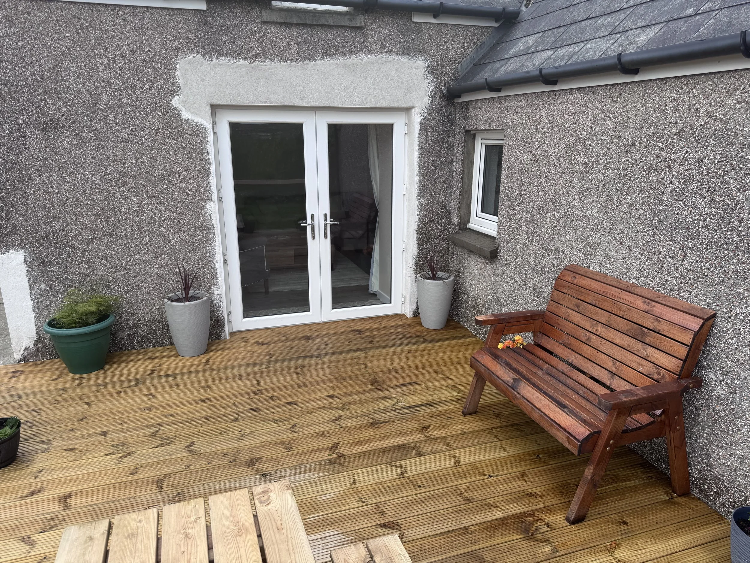 A small outdoor patio area with a wooden deck and a sliding glass door leading inside. There are two large potted plants beside the door, one in a green pot and the other in a white pot. A wooden bench with flowers on it is positioned on the right si