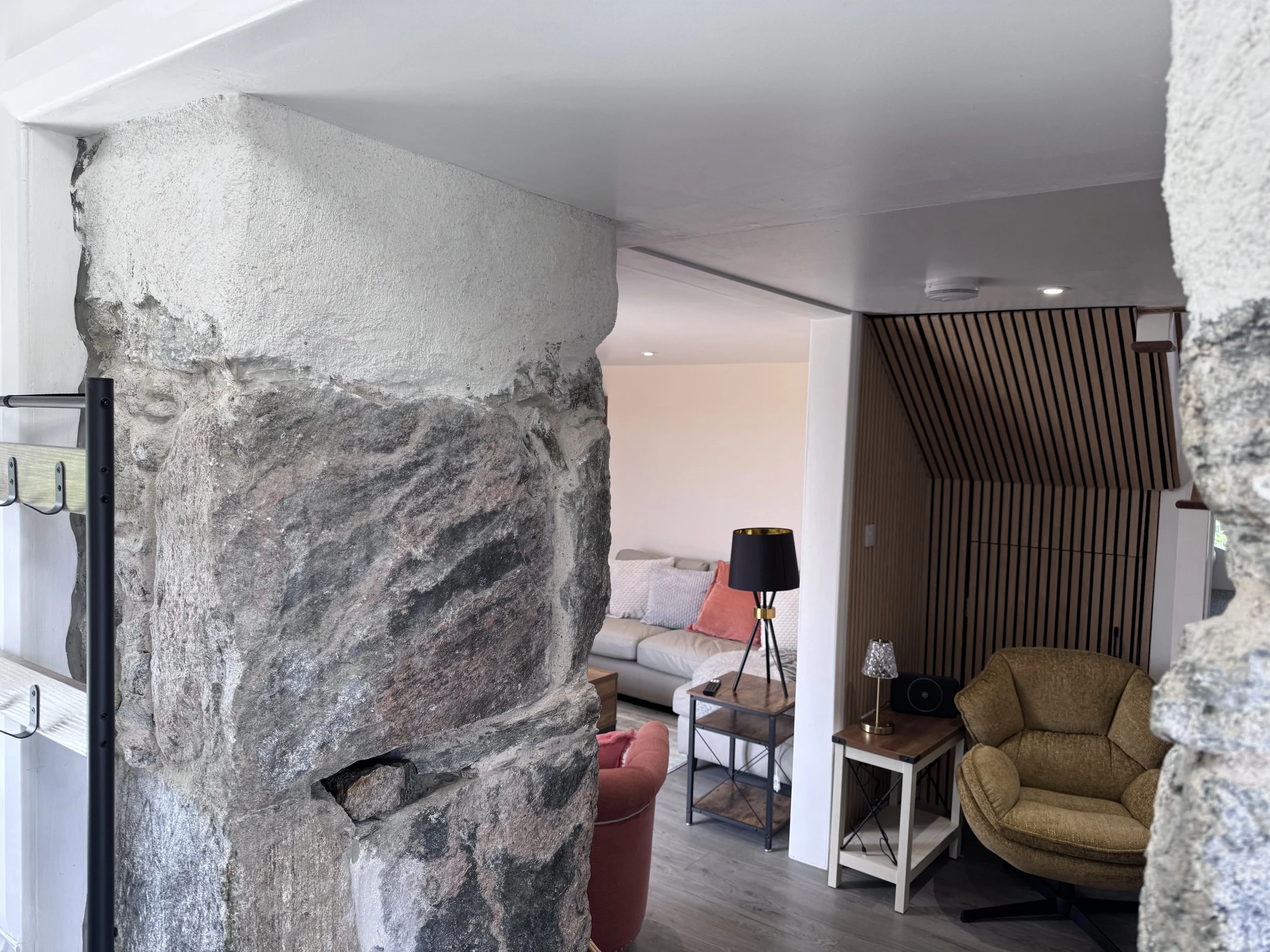 Interior view of a living room with a large rock wall feature, white and wooden accents, a tan armchair, a black and gold lamp, and a window withblinds.