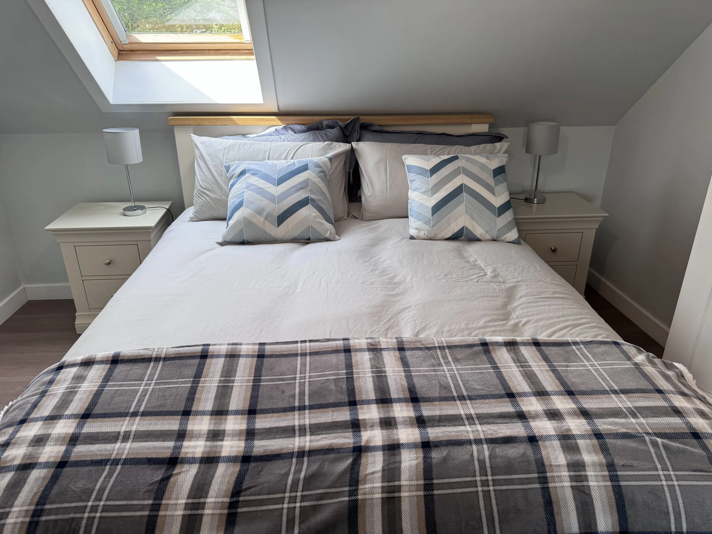 A cozy bedroom with a bed placed against a sloped ceiling under a skylight window. The bed has a white sheet, two white pillows, and two decorative pillows with blue and gray chevron patterns. On each side of the bed are matching white nightstands, each with a white lamp. The room is lit by natural light coming through the skylight.
