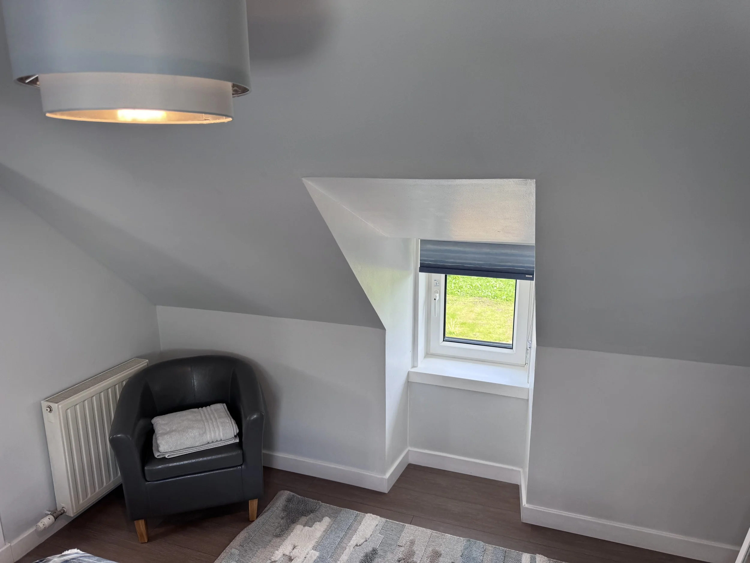 A cozy corner with a black armchair holding a folded white towel, positioned beneath a window with open blinds revealing a grassy yard outside, amidst white walls and dark wooden flooring.