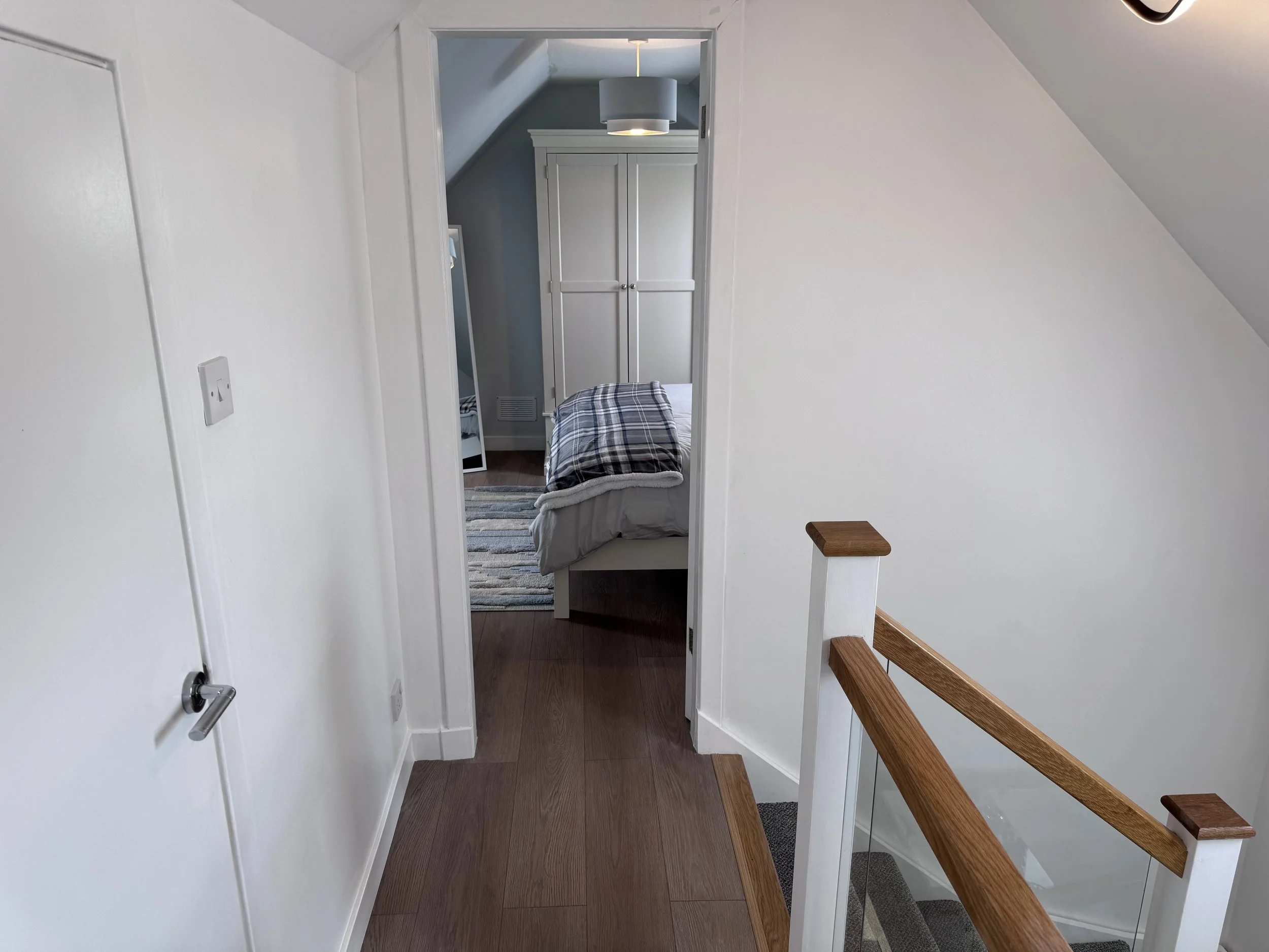 A doorway leading to a bedroom with a bed, plaid bedding, a striped rug, a white wardrobe, and a gray ceiling lamp. White walls and hardwood floors are visible, with a staircase railing on the right.
