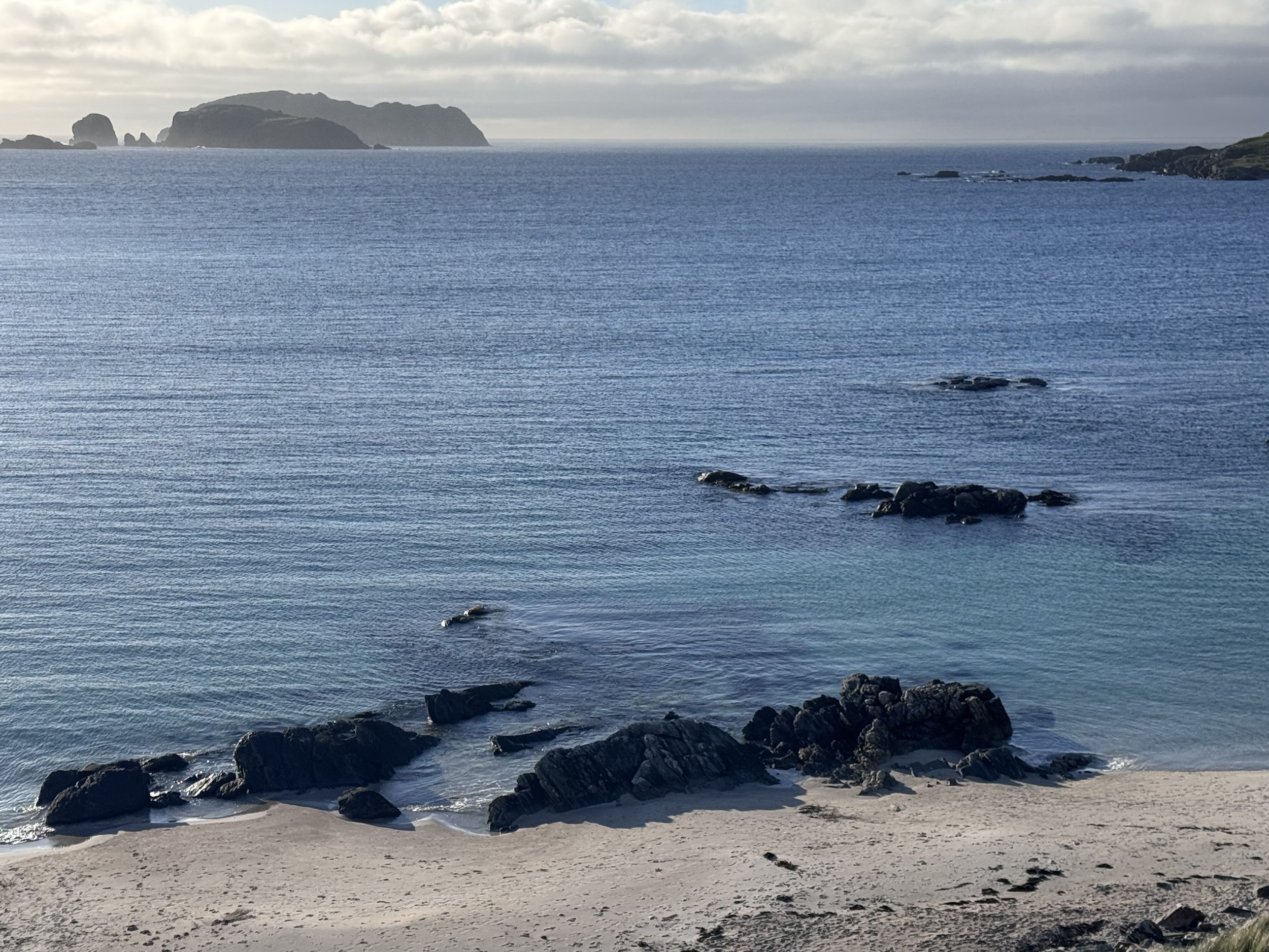 A coastal scene with rocks on the sandy beach in the foreground, calm blue water, and distant islands or landmasses under a cloudy sky.
