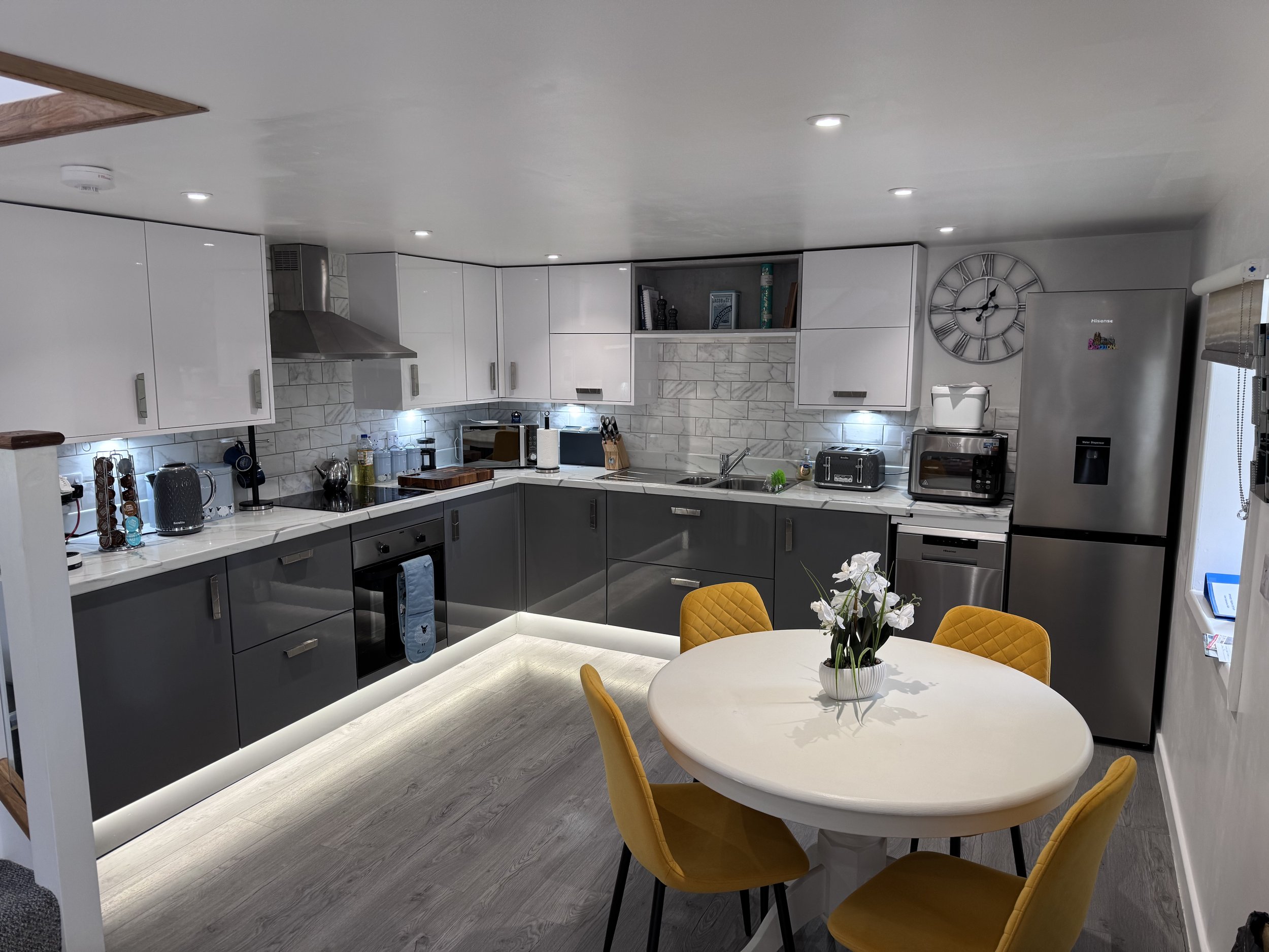 A modern kitchen with white and gray cabinets, marble backsplash, stainless steel appliances, and a round white dining table with yellow chairs.