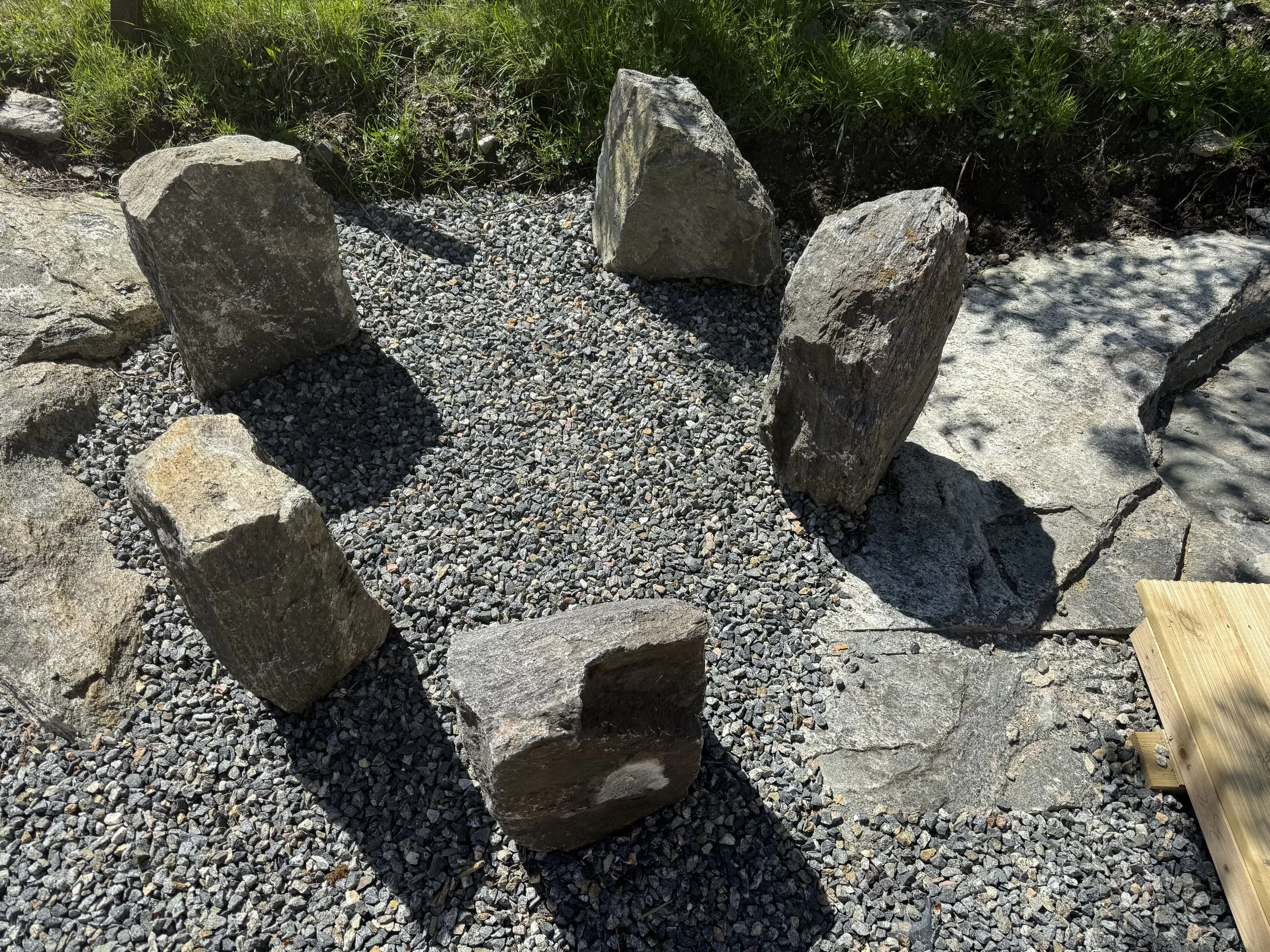 Three large rocks are arranged vertically in a gravel area with grass and soil in the background, and a wooden plank partially visible on the bottom right corner.
