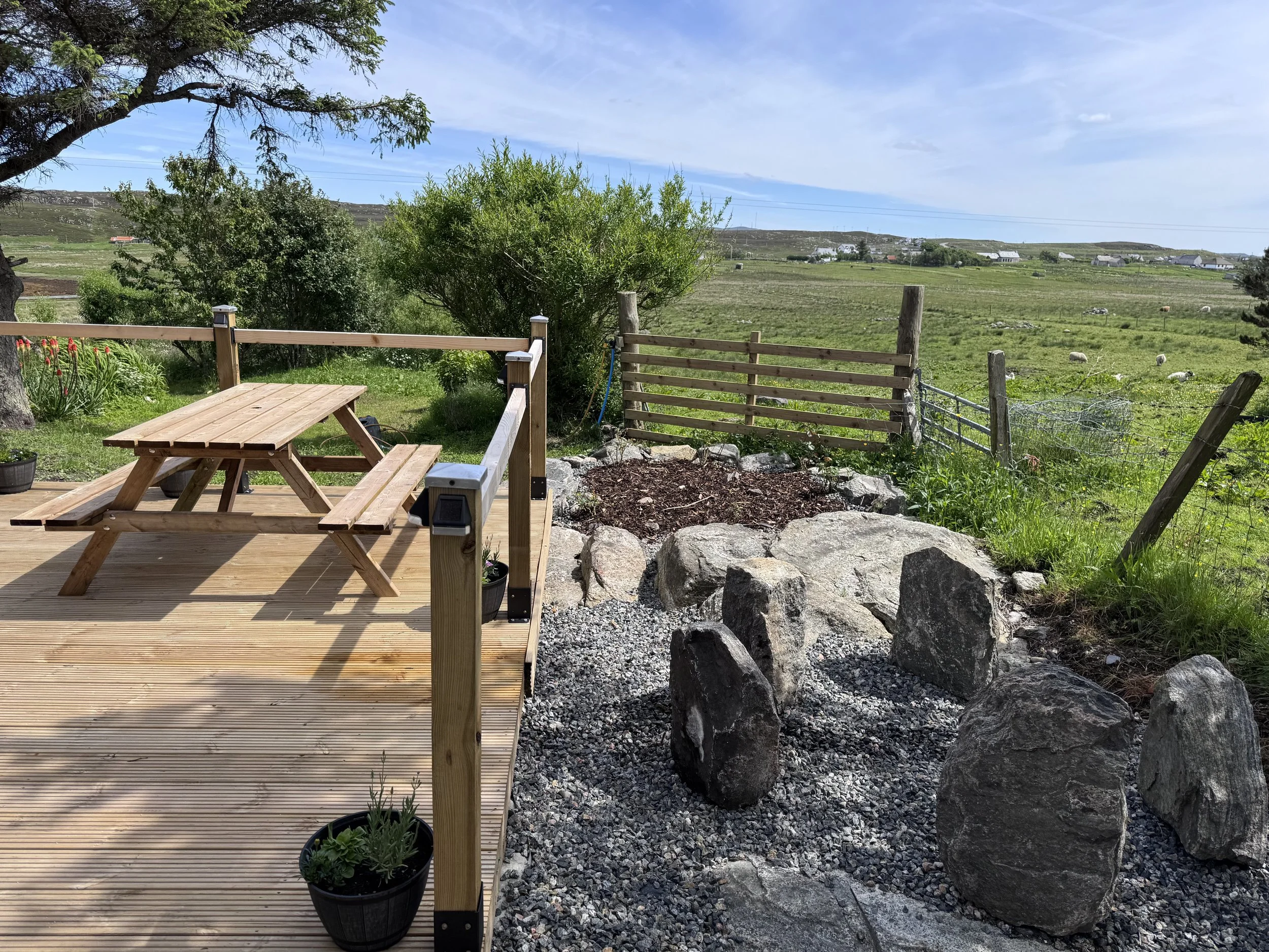 A wooden deck with a picnic table and a potted plant, overlooking green fields with some grazing sheep, trees, and a distant small village under a blue sky with some clouds.