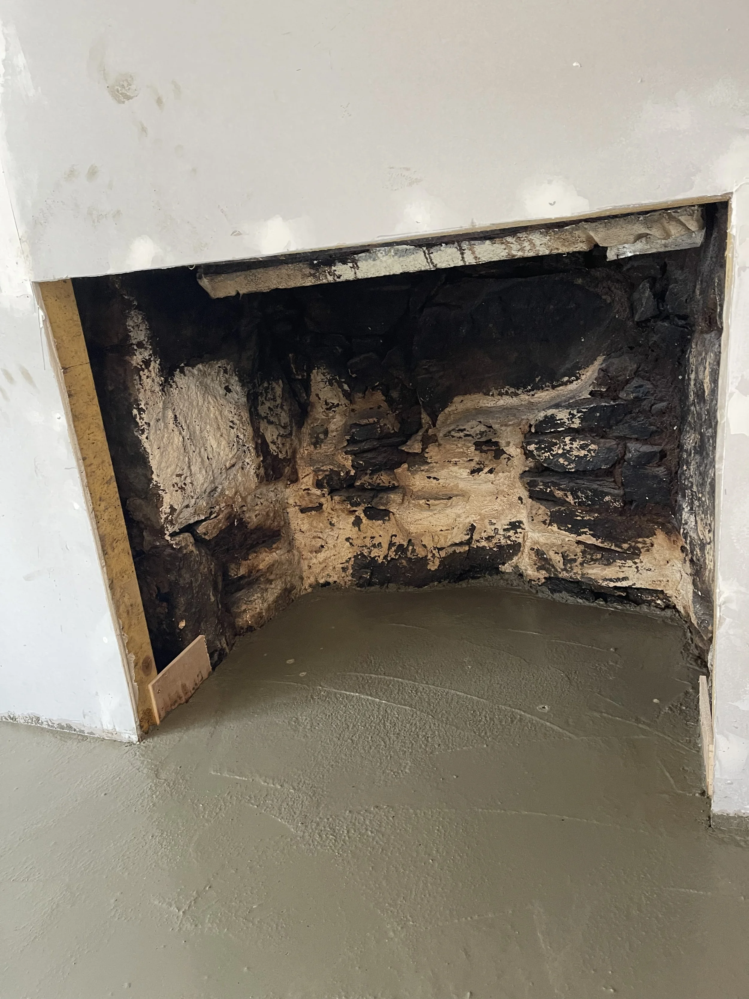 Photo of a construction site showing a doorway opening with a rough, unfinished rock wall inside. The surrounding wall is drywall with some patches and a piece of wood at the bottom left.