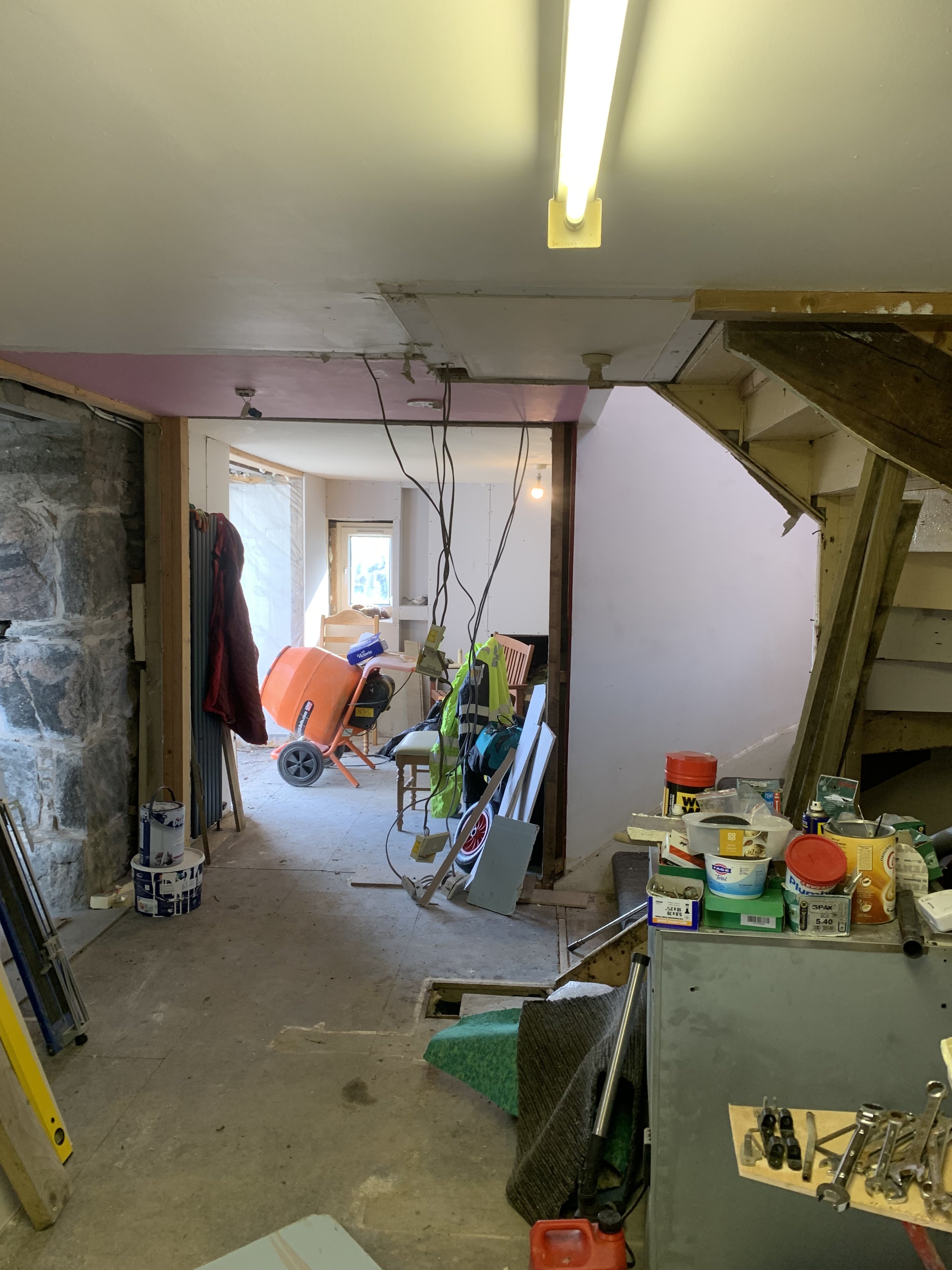Construction or renovation site with unfinished walls, tools, and supplies scattered around. Visible drywall, electrical wires hanging from the ceiling, and a mix of construction materials.