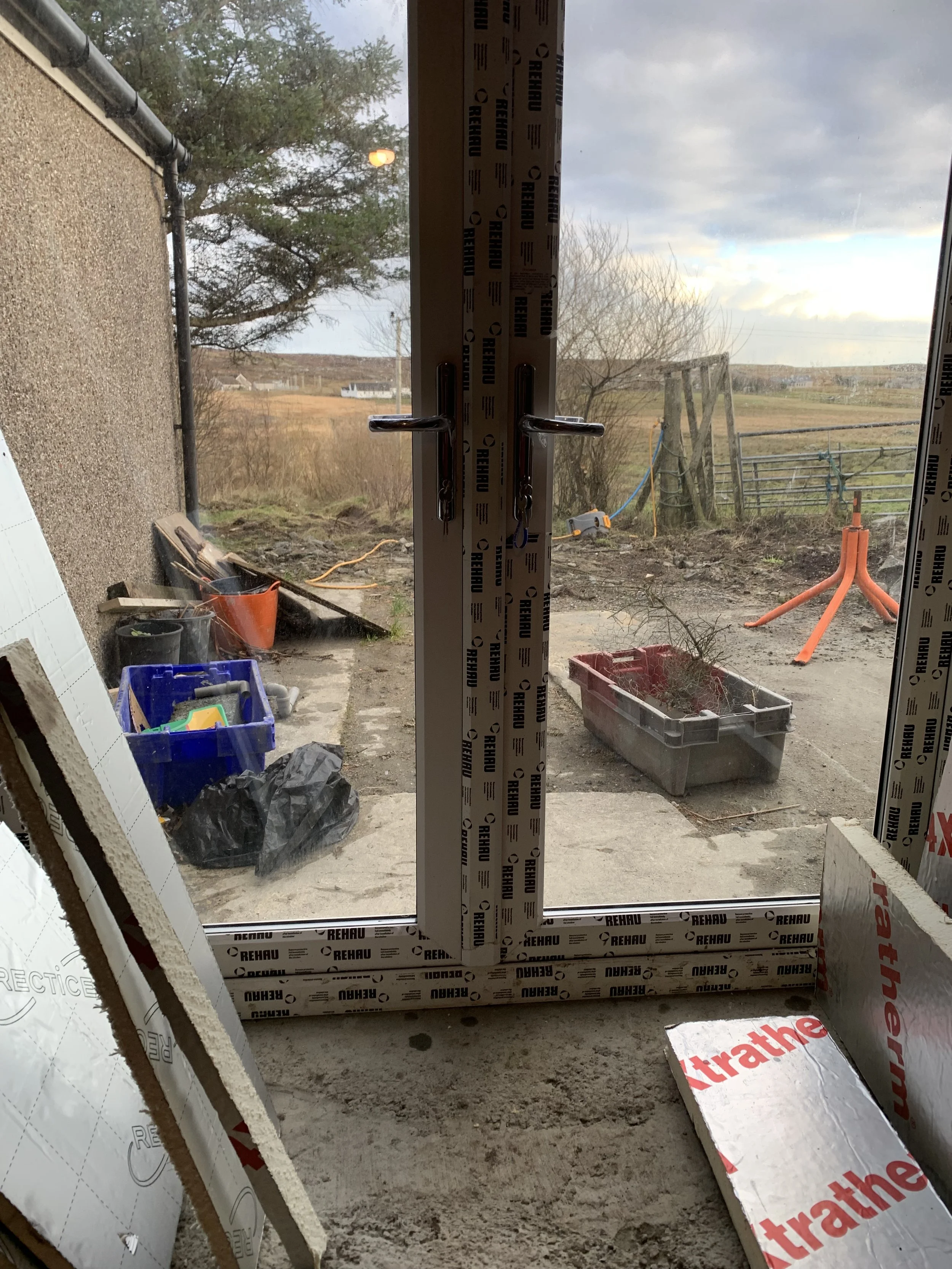 View through a window showing a rural outdoor scene with a cloudy sky, leafless trees, a lawn, a fence, and construction tools and materials outside. Inside are various construction supplies, including a box labeled 'Thrathe' and black trash bags.
