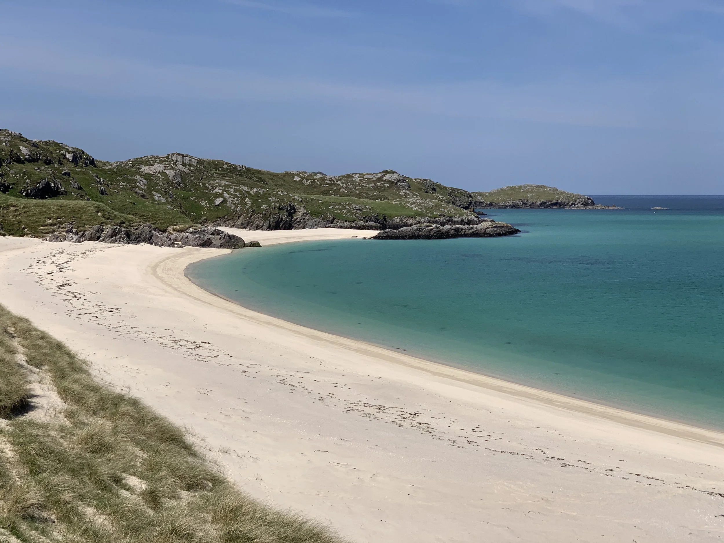 A scenic view of a sandy beach with turquoise water, rocky cliffs, and green grass-covered hills under a partly cloudy sky.