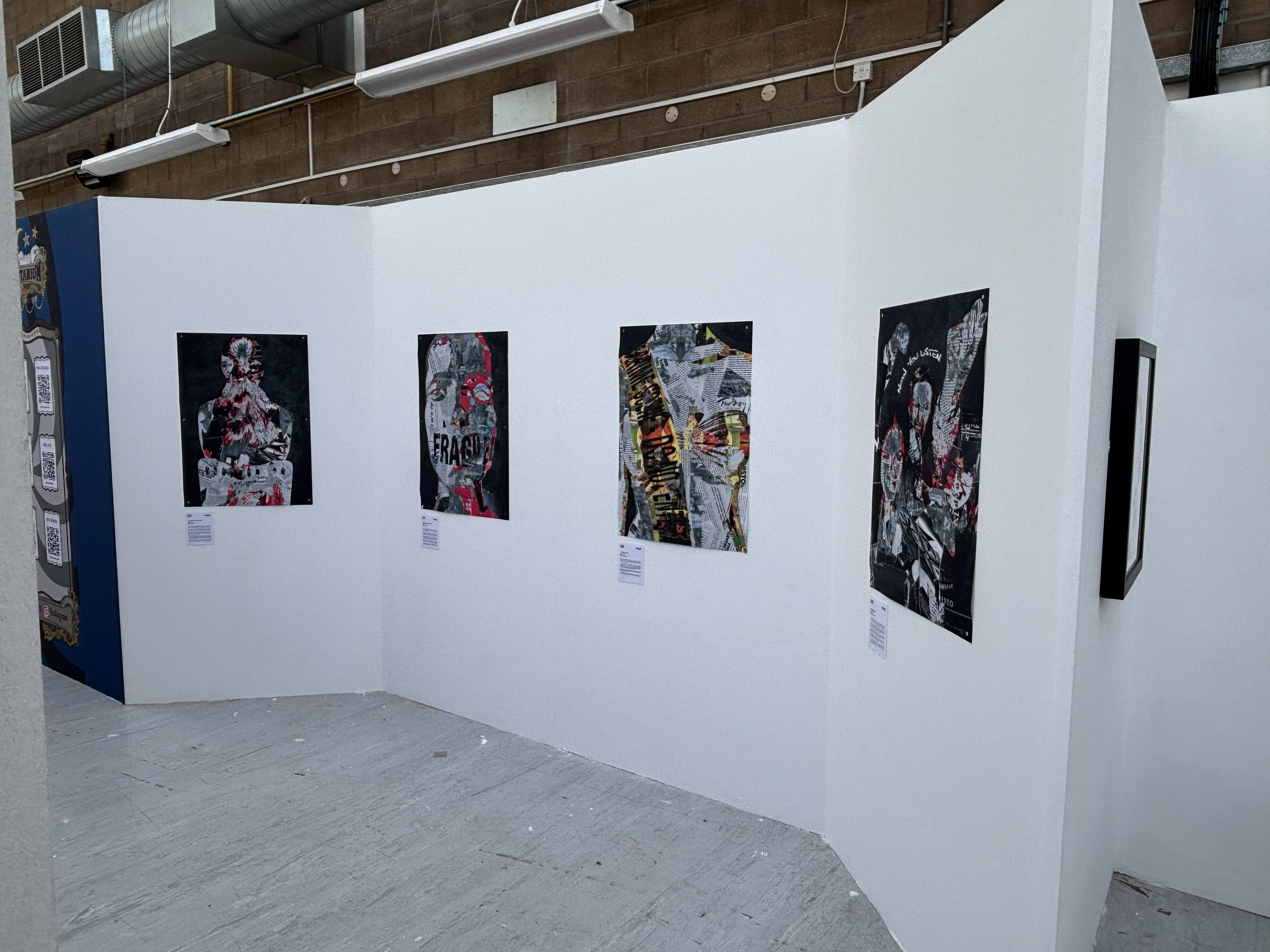 Art exhibit featuring posters with collages and text on a white partition wall in an industrial-style gallery space.
