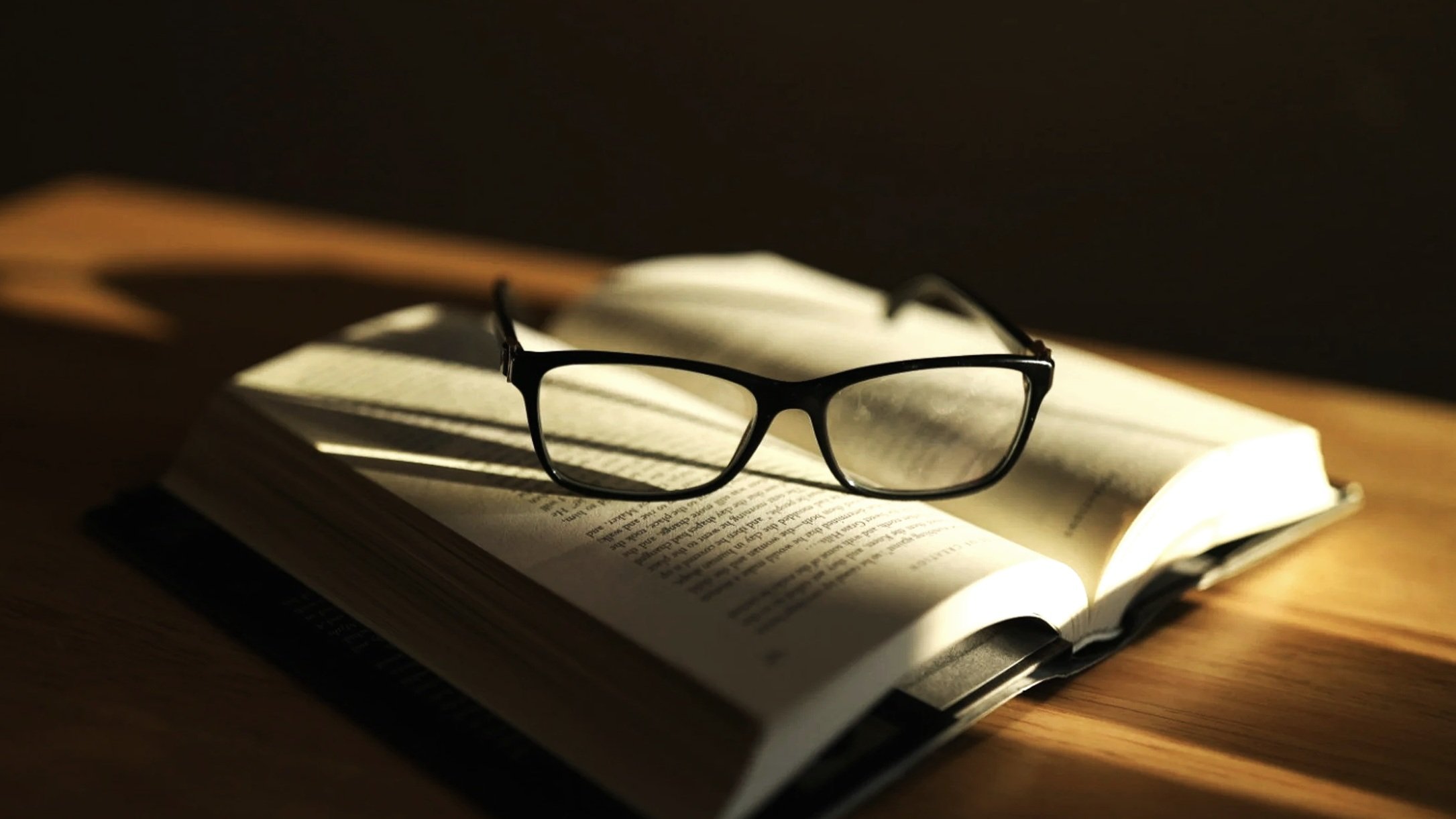 A pair of black eyeglasses resting on an open book with reading glasses casting shadows in warm lighting.