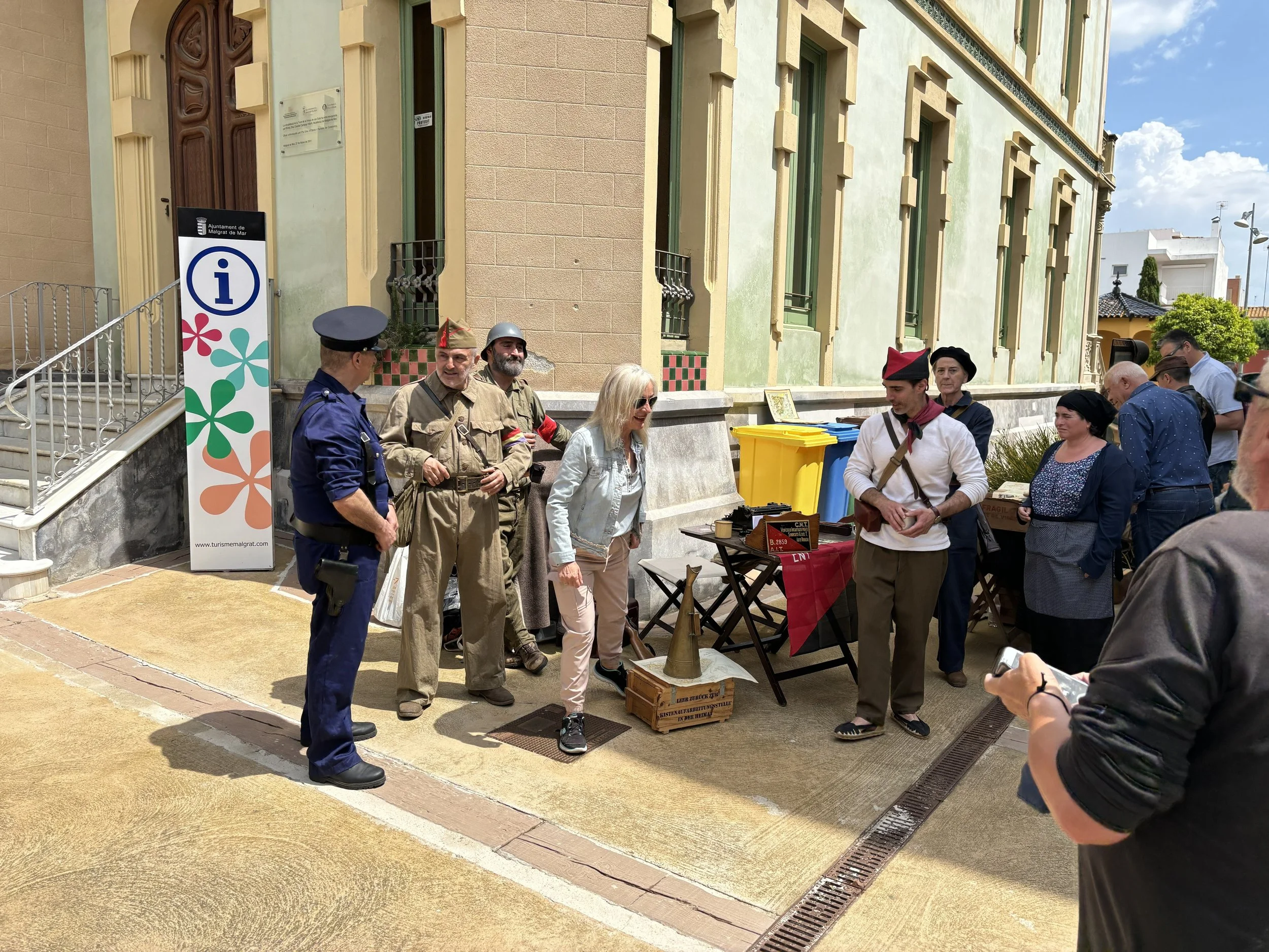 People dressed in historical military uniforms and civilian clothing standing outside a building with a sign indicating information. There are colorful trash bins and a display of historical items. The scene appears to be a historical reenactment or outdoor event.