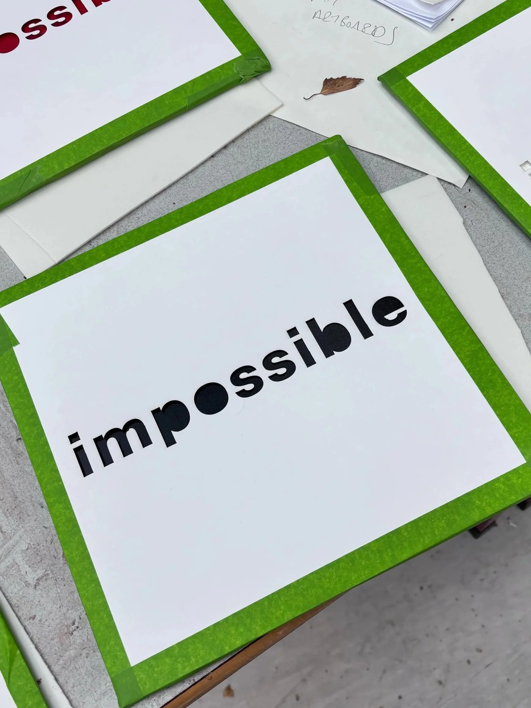 A white paper with the word 'impossible' printed on it, bordered in green, lies on a gray surface with a small dry leaf nearby.