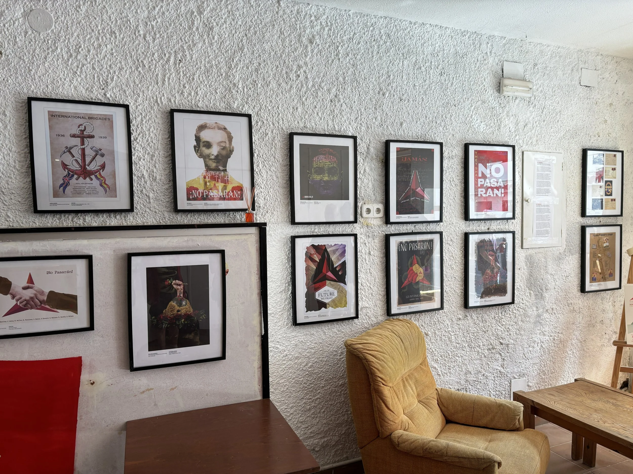 A textured white wall with a collection of framed posters and images, including revolutionary and political themes, with a yellow armchair nearby and a small wooden side table.
