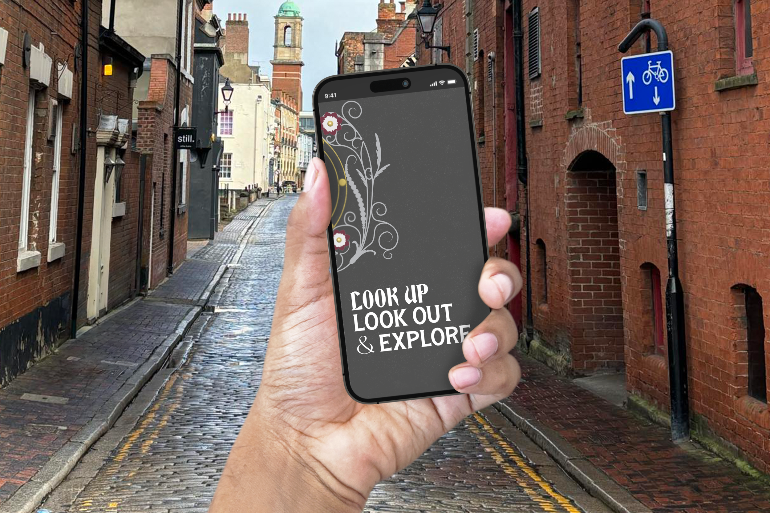 A person holding a smartphone with a decorative screen displaying the text "LOOK UP, LOOK OUT, & EXPLORE". The background shows a cobblestone street lined with brick buildings and a blue bicycle lane sign.