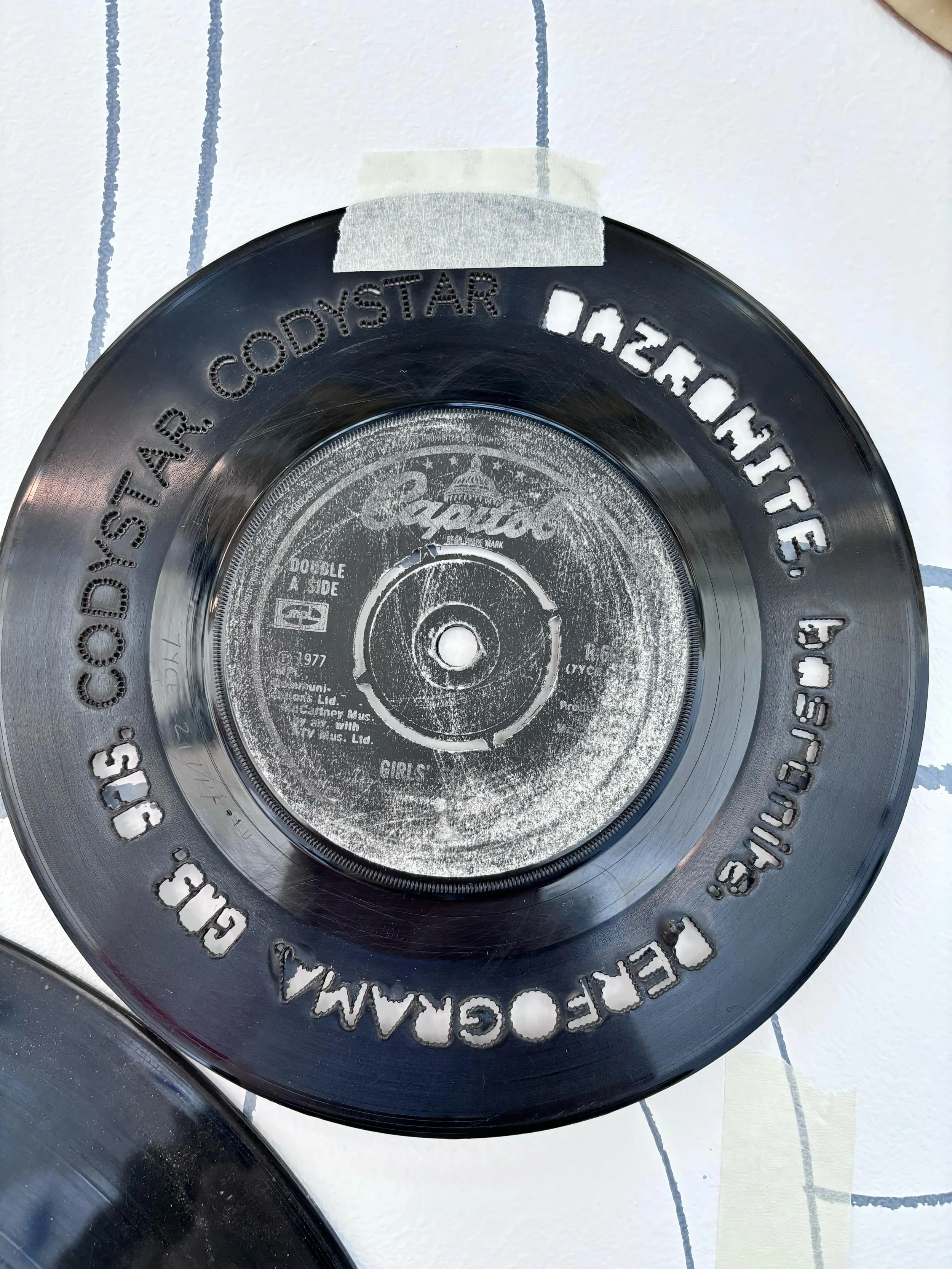 Close-up of a vinyl record with a tape on the side and detailing around the edge, placed on a tiled floor.