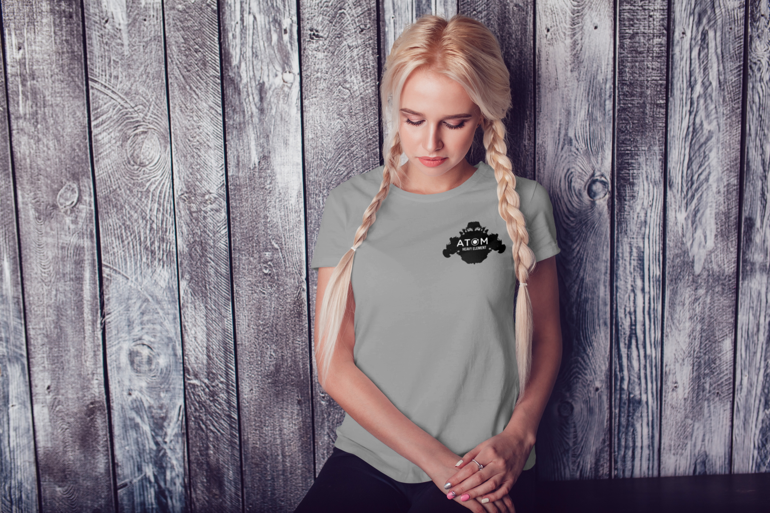 A young blonde woman with braided hair standing in front of a rustic wooden wall, wearing a gray T-shirt with a black cloud logo and the words "ATOM Heavy Element." She has her eyes closed and her hands clasped at her waist.
