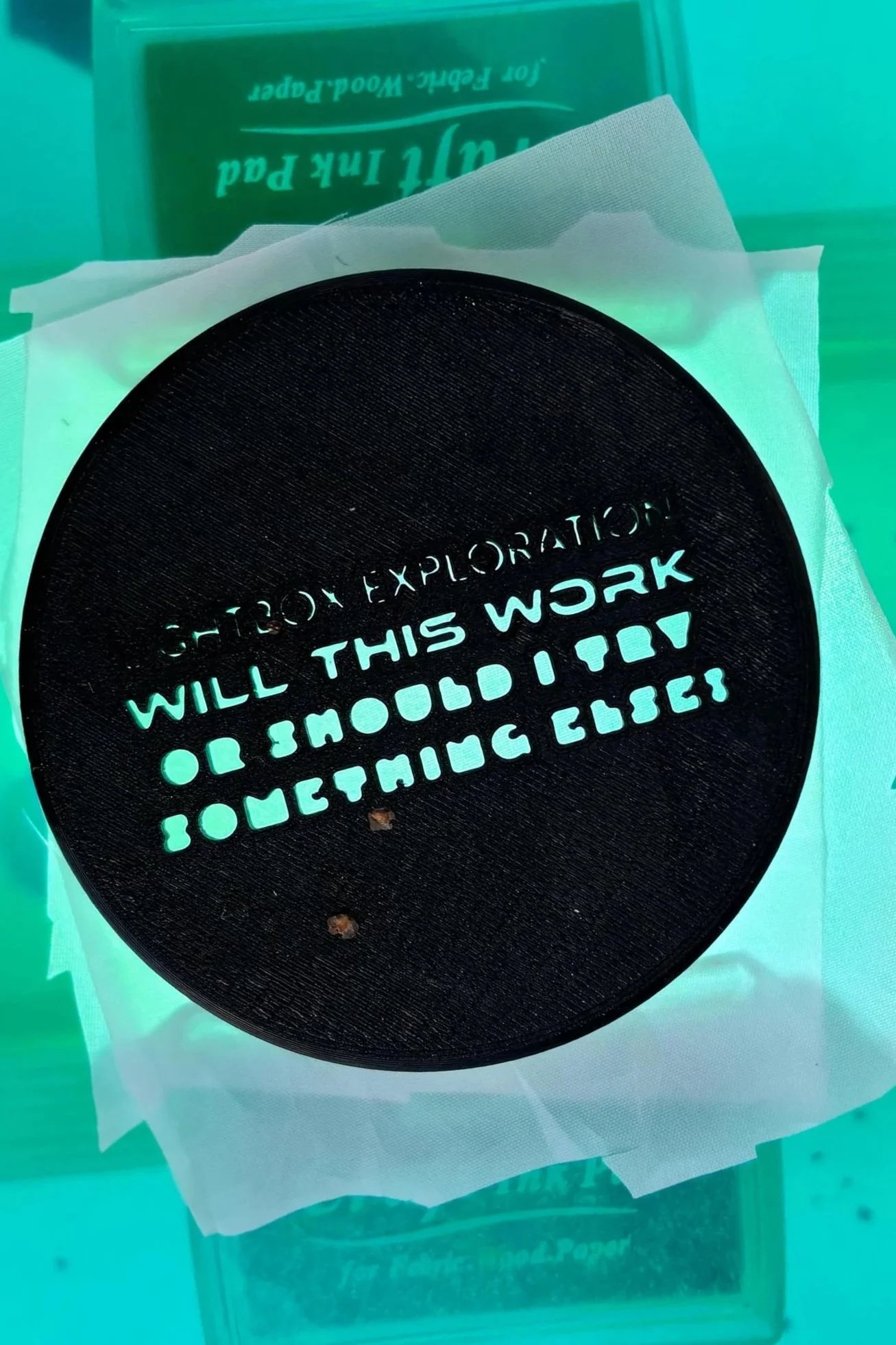 Close-up of a circular black object with green text that reads, 'Will this work or should I just do it myself?' It appears to be resting on a white paper or tissue, with a green background and some packaging or box in the blurry background.