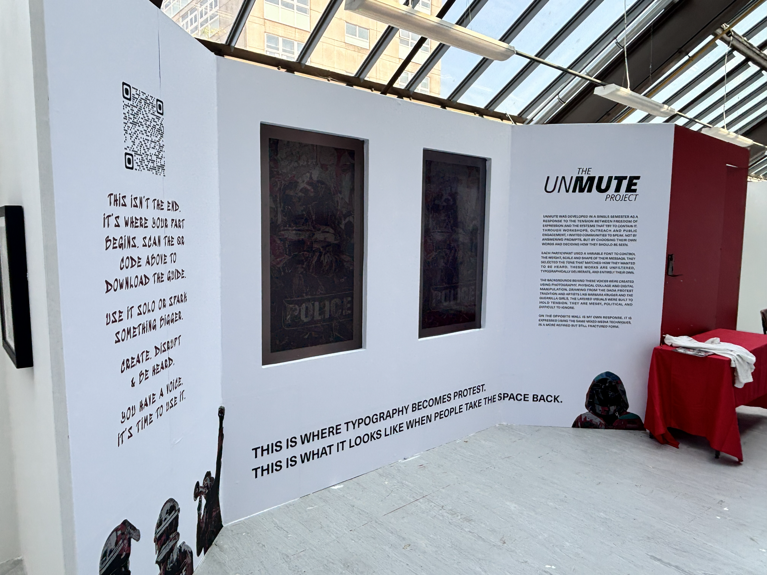 An informational display panel titled "The Unmute Project" with text about the project's development, activities, and dedication to community engagement. The panel includes a QR code, two rectangular windows, and a red table with white cloth and papers nearby, set inside a building with a glass ceiling and city buildings in the background.