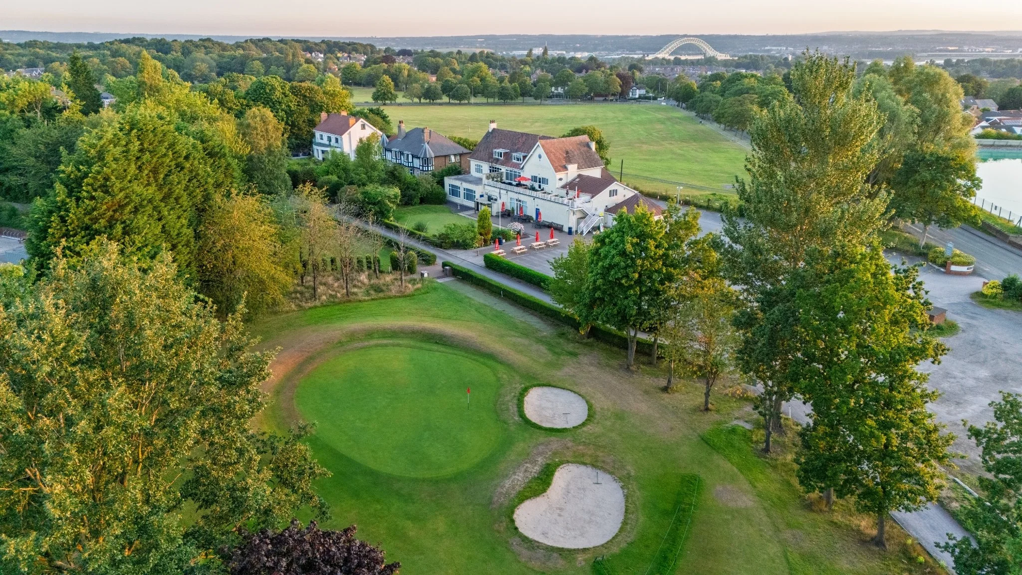 Aerial view of a golf course with green grass, sand bunkers, trees, a parking lot, residential houses, a large park, and a bridge in the distance.