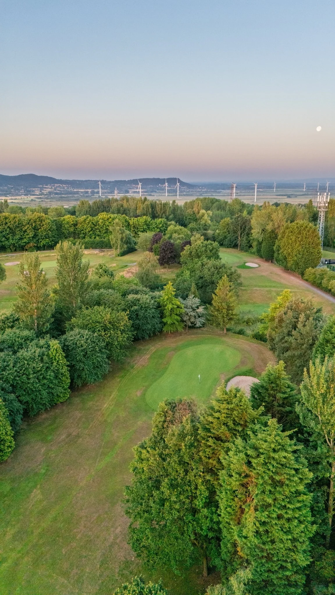 A scenic landscape with a golf course surrounded by trees in the foreground, a flat valley with wind turbines in the middle ground, and hills with a moon in the sky at sunset or sunrise.