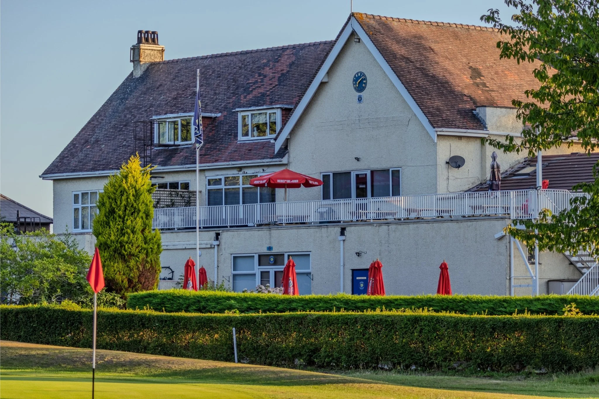 A multi-story house with a red tile roof, a large deck with red umbrellas, green landscaping, and a flagpole in front, situated near a golf course green.