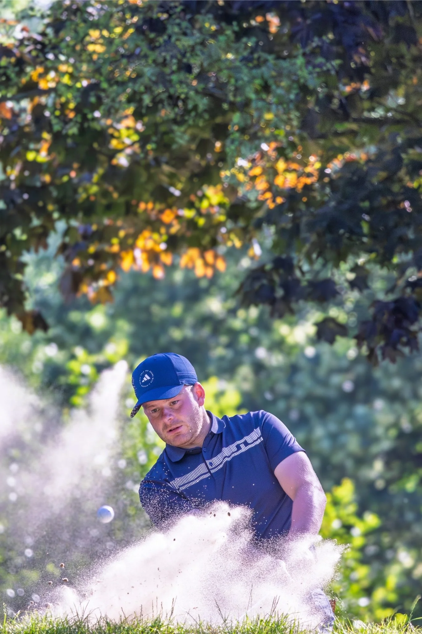 A man in a blue cap and navy shirt hits a golf ball with a club on a sunny day, causing a cloud of dust to spray up from the ground with green trees and foliage in the background.