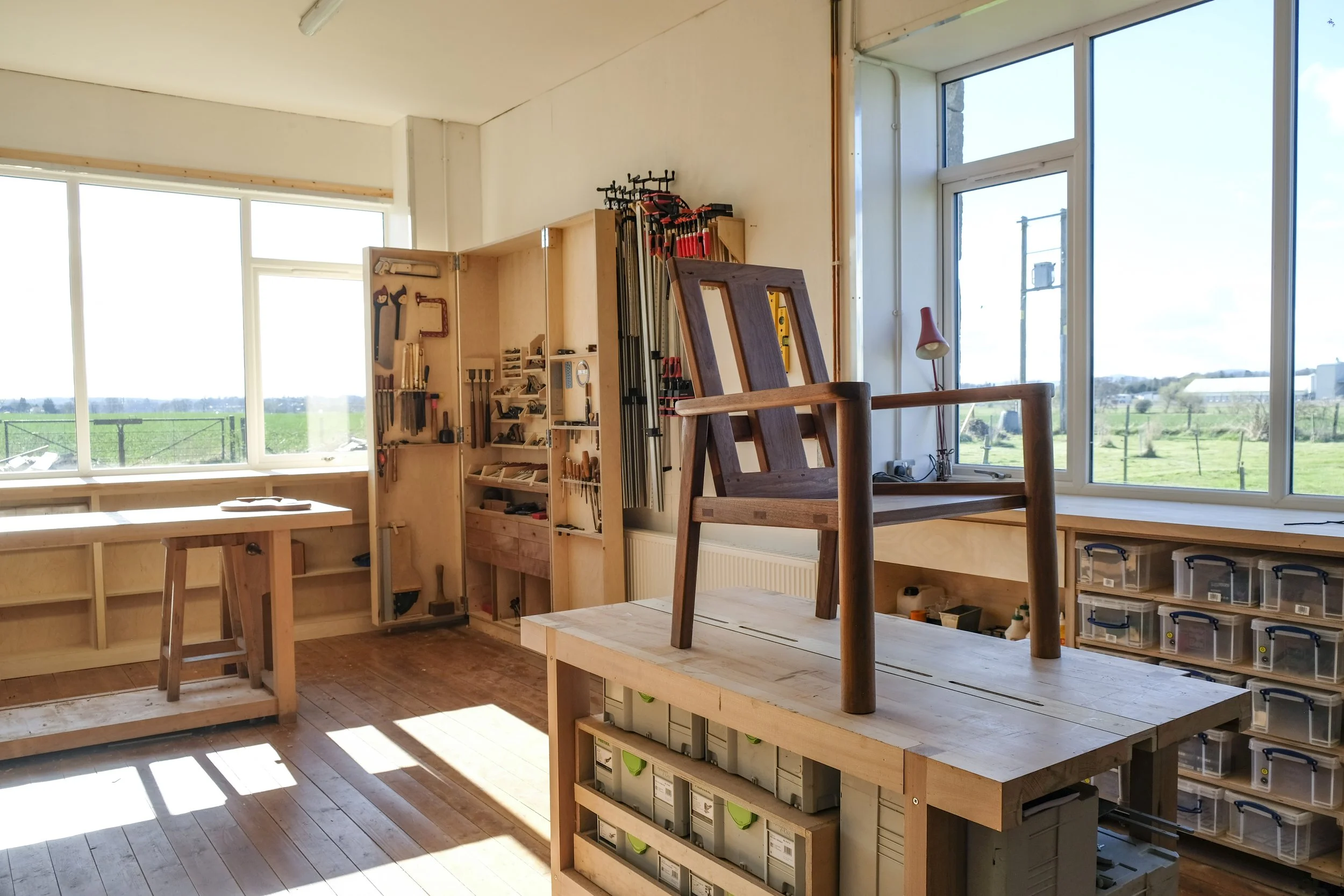 Interior of a woodworking studio at Adam Stone Furniture with large windows, workbenches, a wooden chair in progress, and various tools and materials organized on shelves.