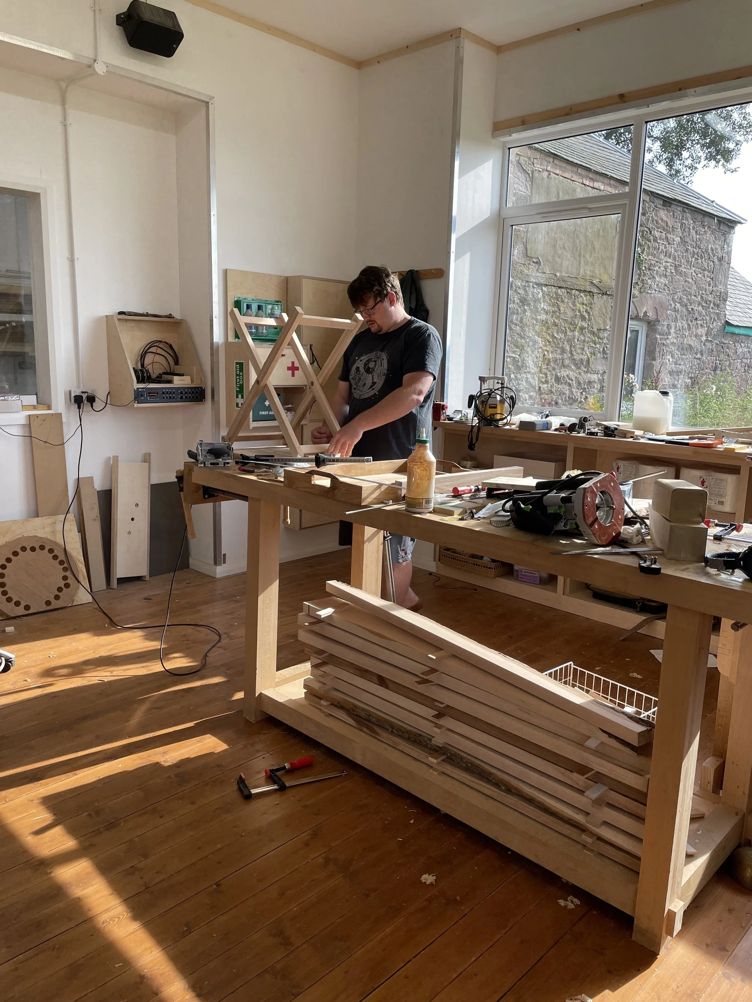 A man working on a woodworking project in a bright workshop, surrounded by tools and wood pieces, with a large window letting in natural light at Adam Stone Furniture.