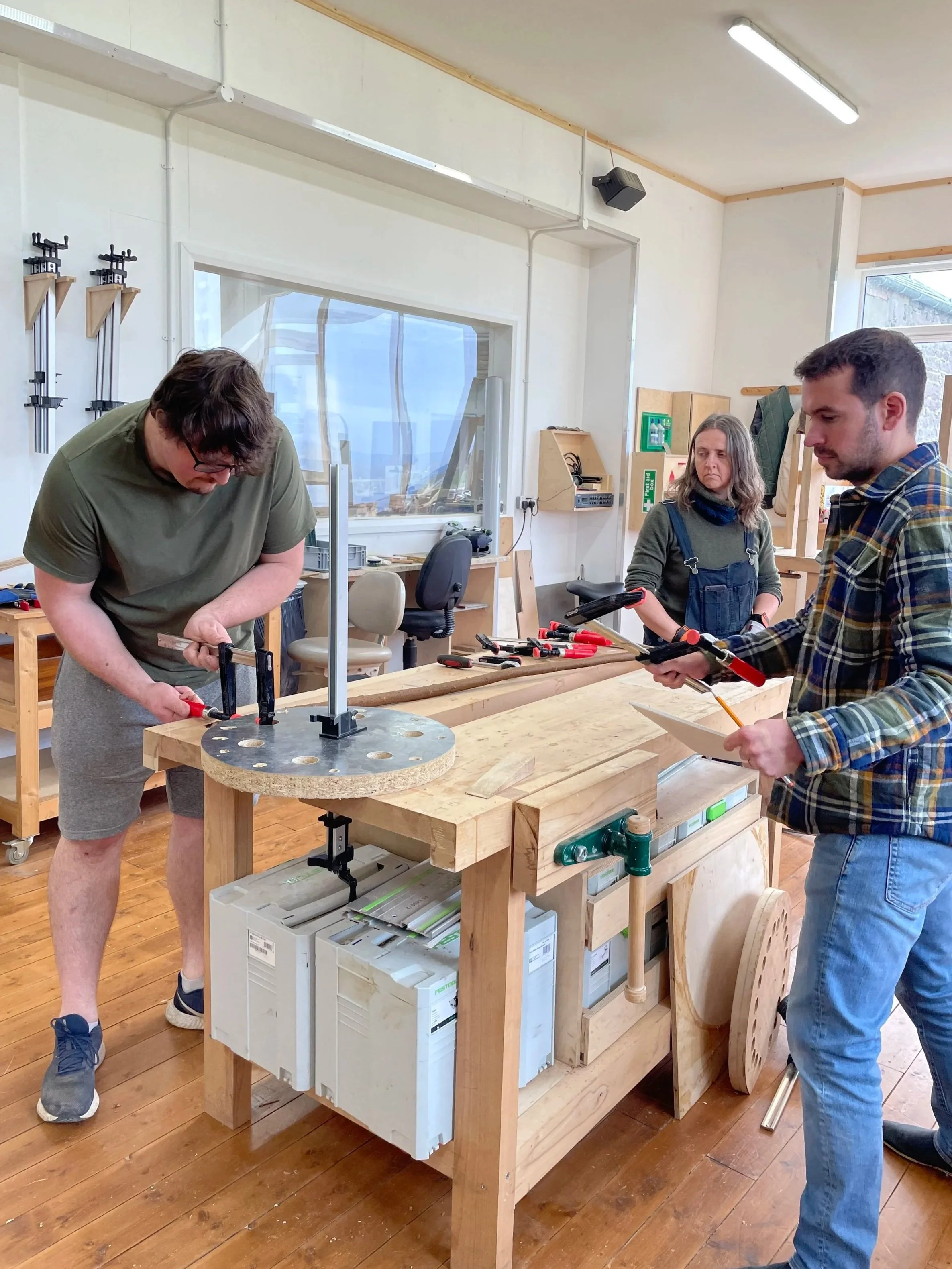 Three people working together on a woodworking project in a workshop. They are using clamps and power tools on a wooden workbench, with shelves and workshop equipment visible in the background at Adam Stone Furniture.