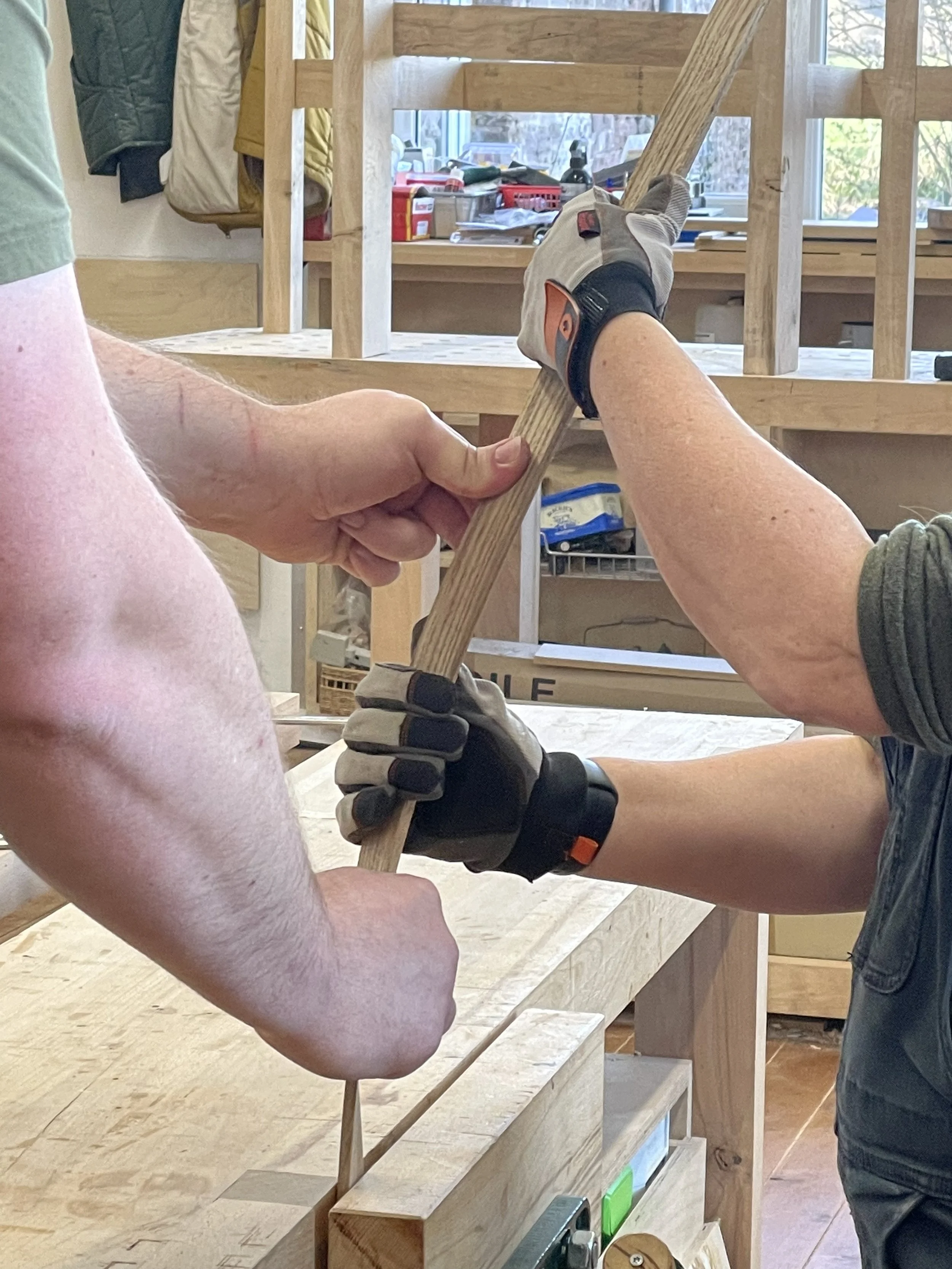 A person wearing a work glove, bending steamed ash strips by hand in a woodworking shop at Adam Stone Furniture.