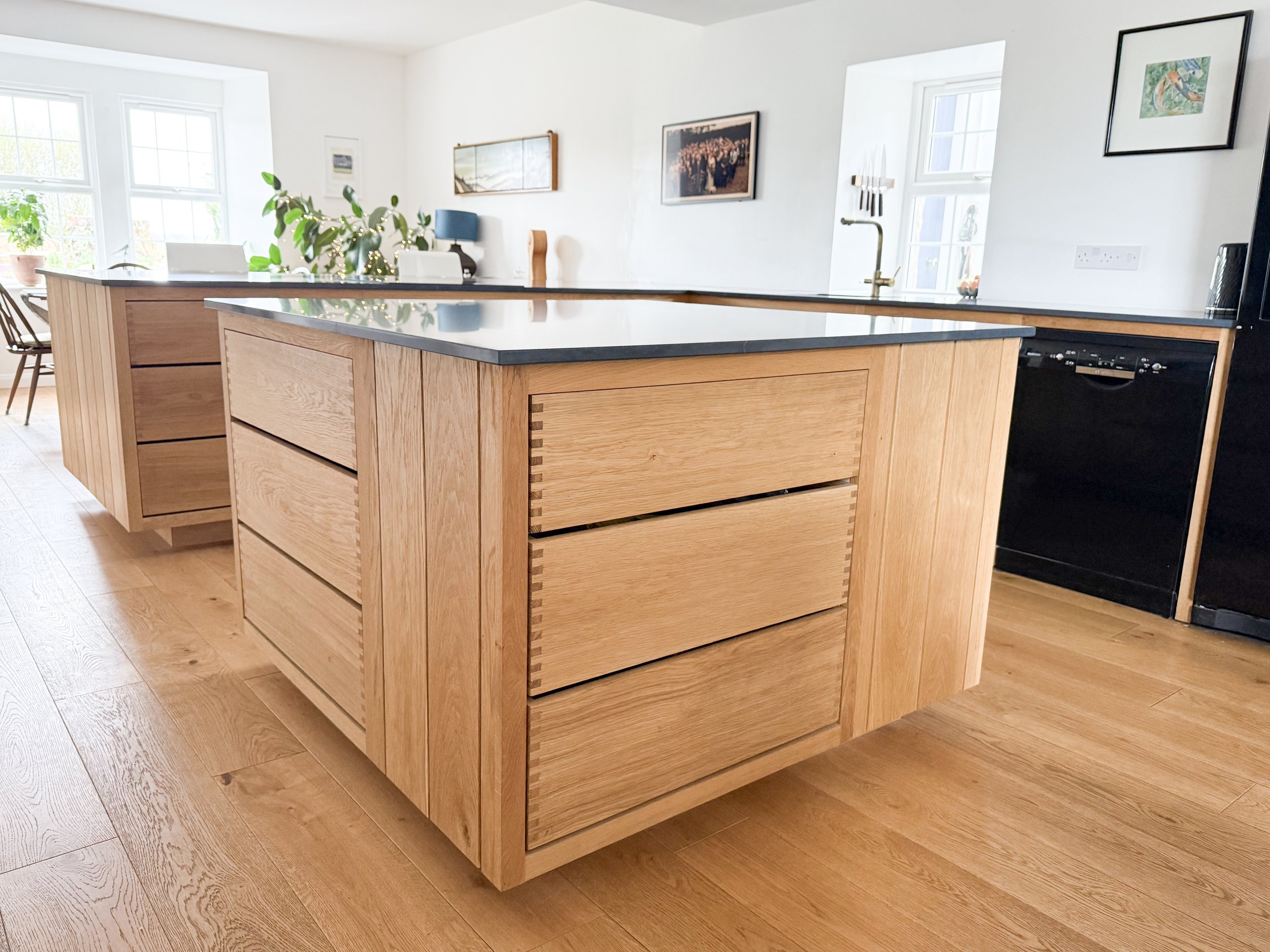 A modern oak, finger-jointed kitchen with a wooden island, Silestone countertop, and framed artwork on the white walls, with bright natural light coming from large windows at Adam Stone Furniture.