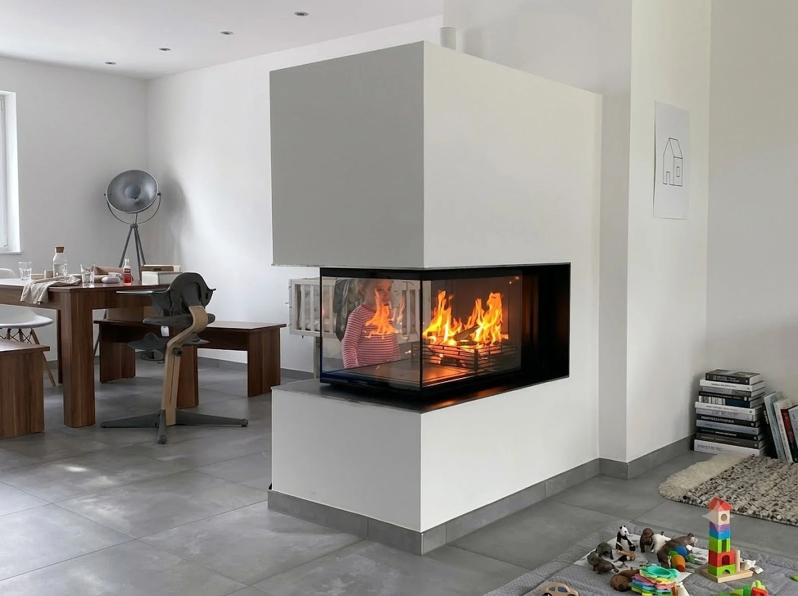 ein im raum integrierter, weisser gaskamin mit feuer drin. vor dem kamin steht ein kind und schaut in die flammen