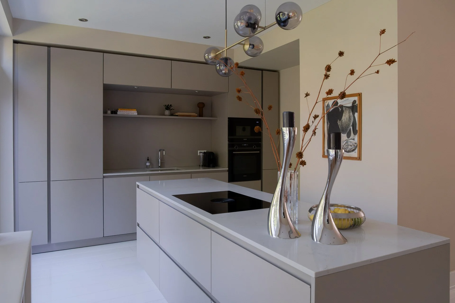 Modern kitchen with beige cabinets, white countertops, decorated with a glass vase containing dried branches with small leaves, a yellow bowl with fruit, and a framed black and white artwork on the wall.