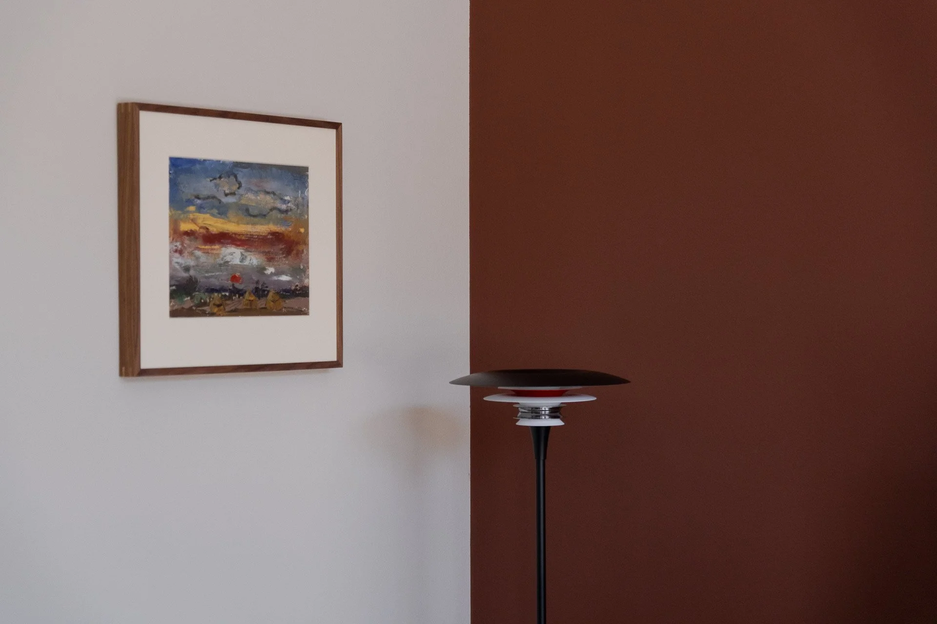 A dual-tone interior room with a white wall on the left and a dark reddish-brown wall on the right. A colorful landscape painting with a wooden frame hangs on the white wall, and a modern black and silver floor lamp with a bowl-shaped shade stands near the corner.