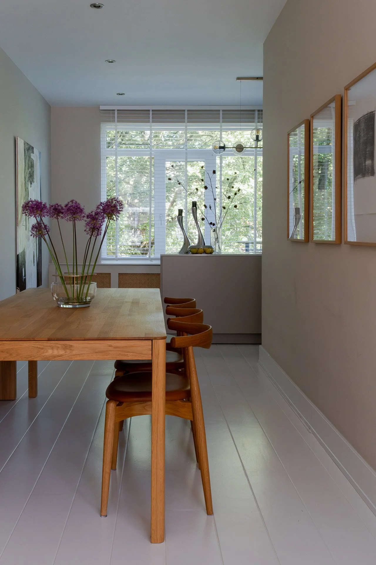 A dining room with a wooden table, four chairs, and a glass vase with purple flowers on the table. Large windows with blinds allow natural light into the room, and decorative vases are on a console beneath the windows. Wall art and mirrors are on the walls.