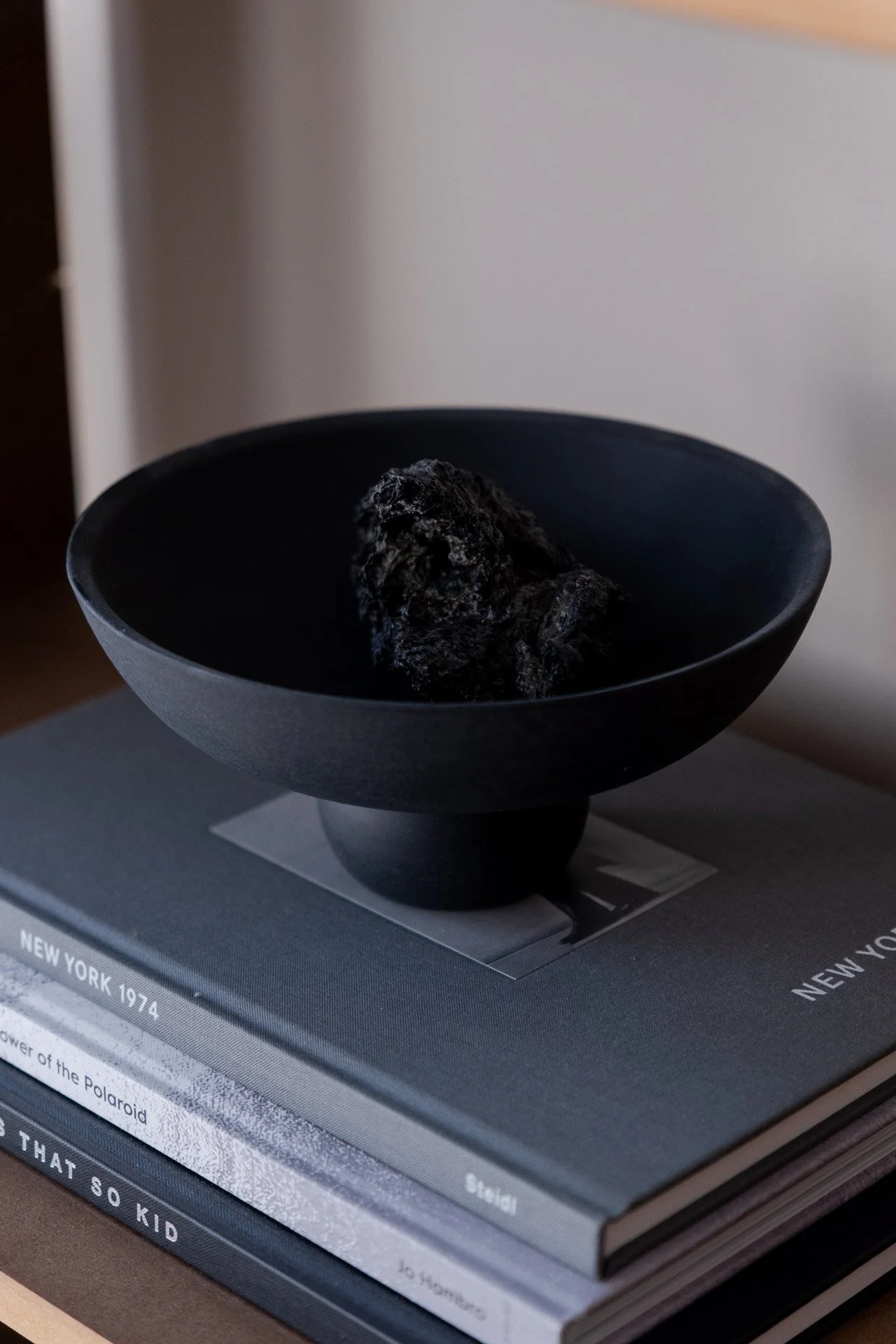 A black bowl with a dark, textured rock inside sits on top of a stack of large books, with a focus on the books' titles and pages. The background is blurred.