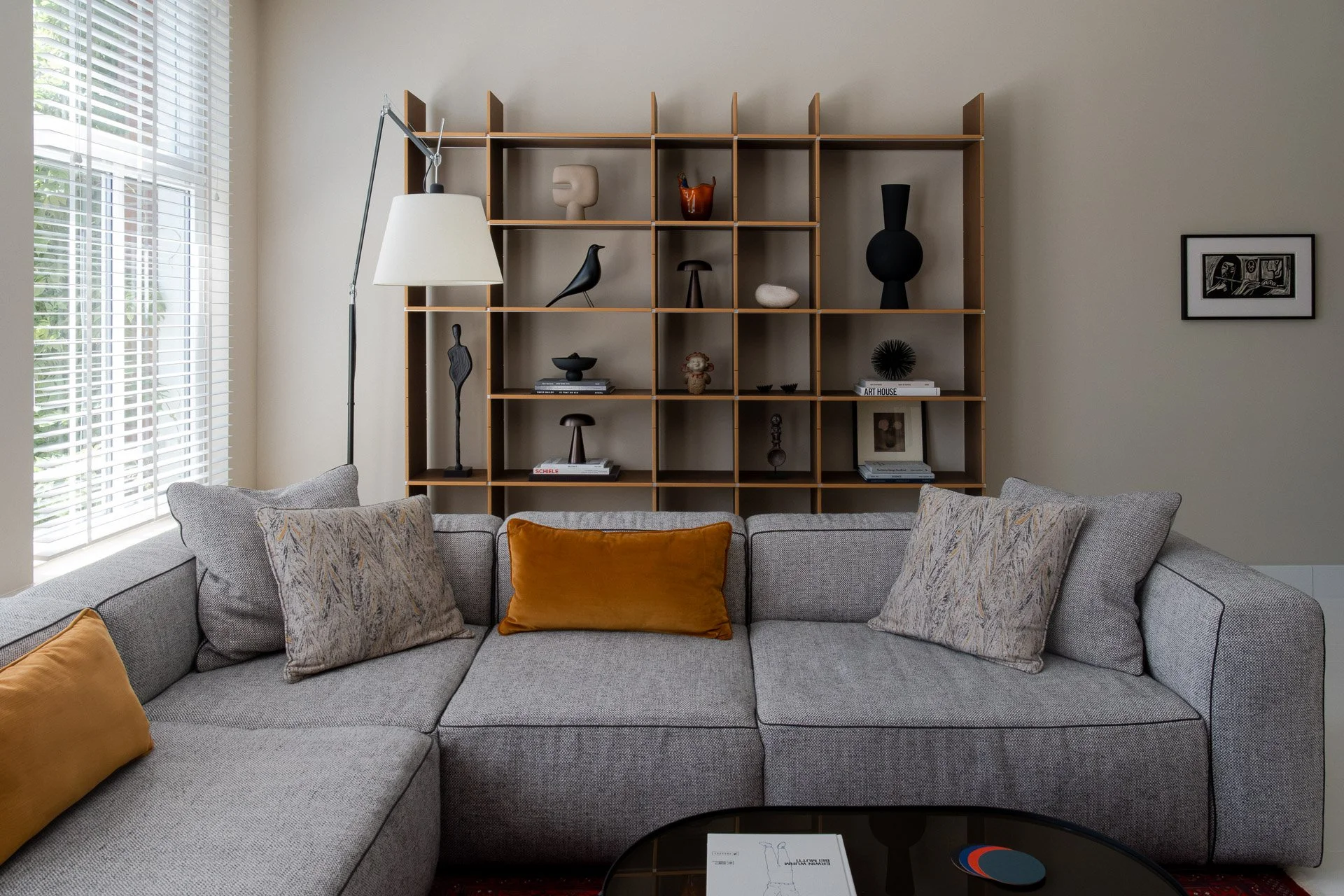 Living room with light gray sectional sofa decorated with patterned and solid throw pillows, a black coffee table, and a wooden shelving unit with art pieces and decorative objects.