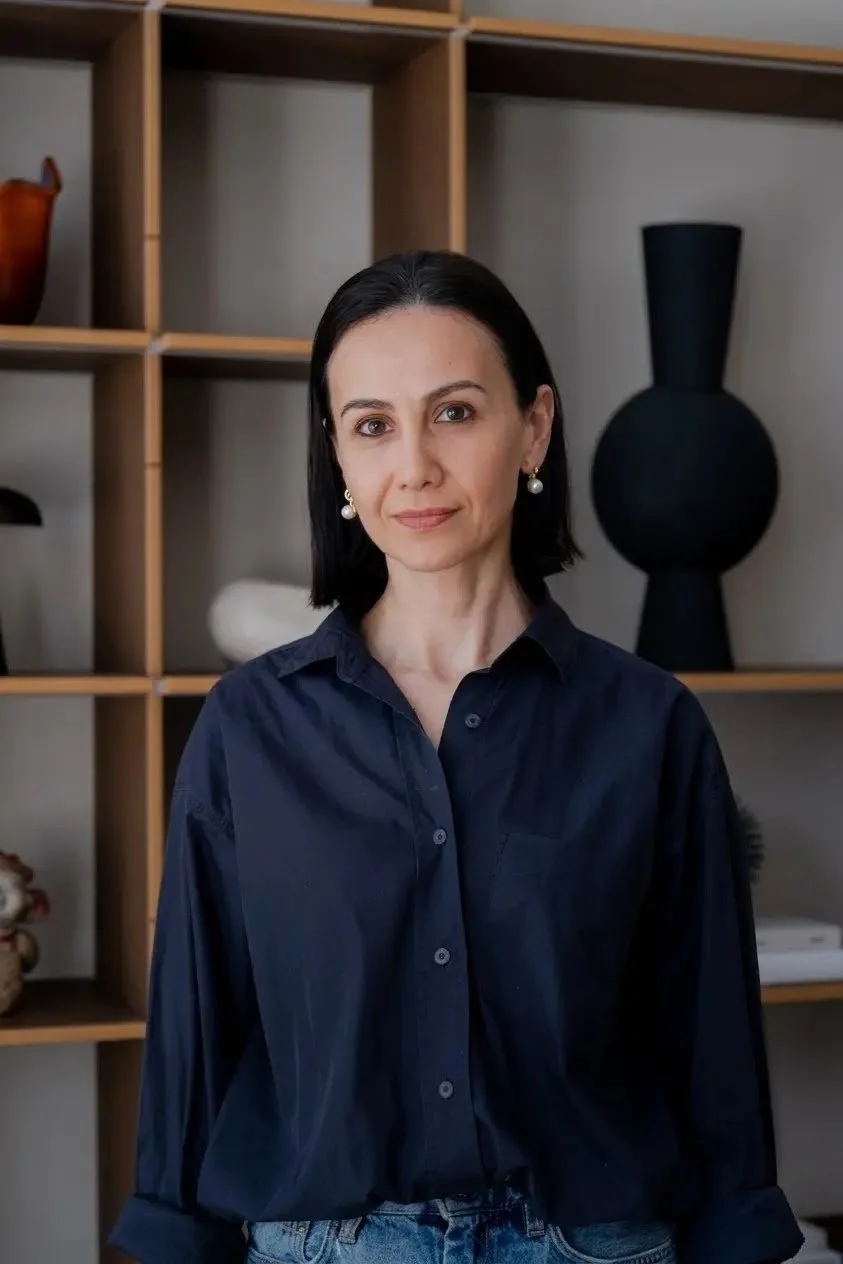A woman with black hair wearing a dark blue shirt and pearl earrings standing in front of a wooden shelving unit with decorative vases and objects.
