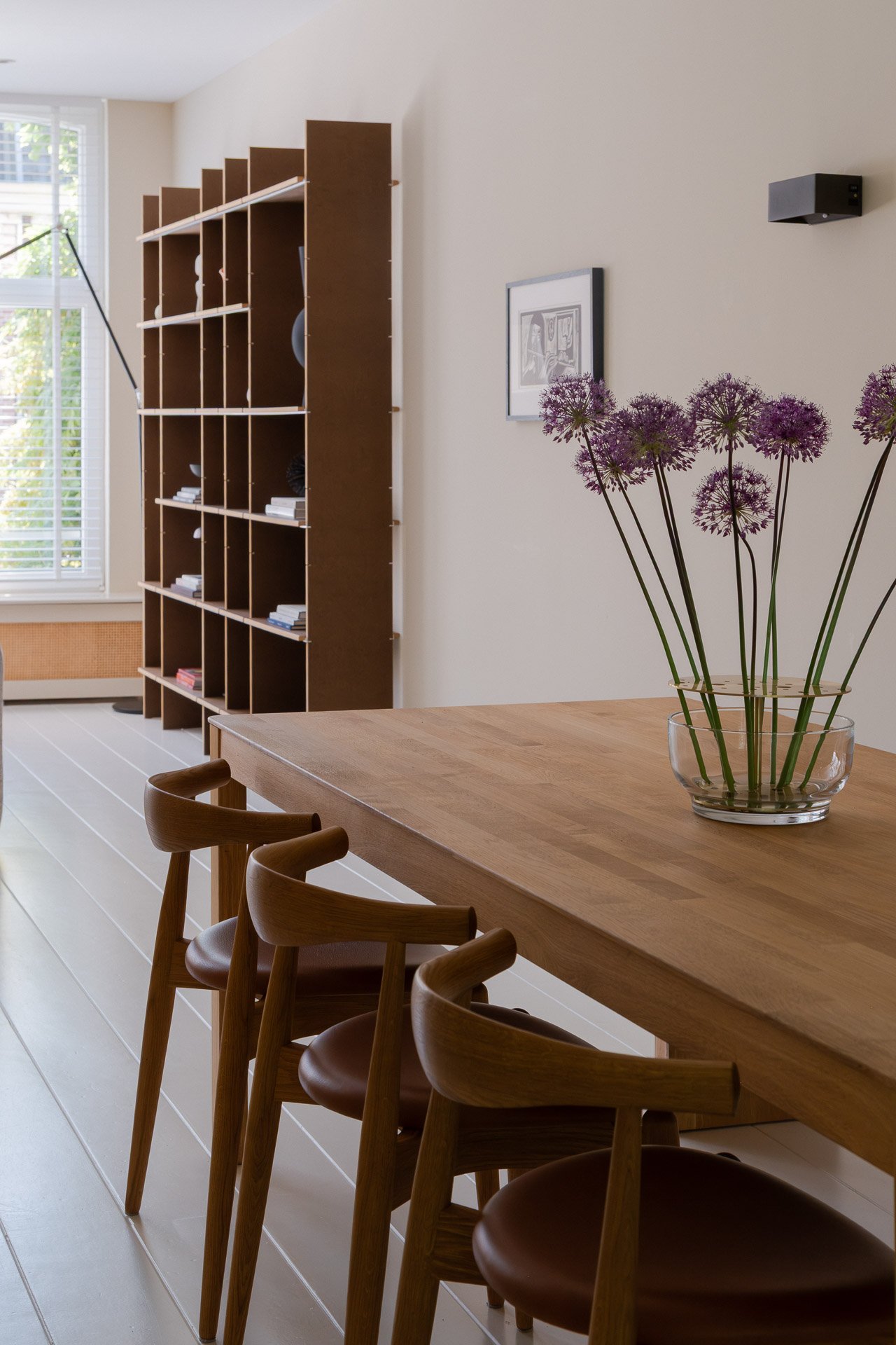 A dining room with a wooden table, six wooden chairs, a glass bowl with purple flowers, a framed black-and-white drawing on the wall, a dark speaker, a wooden bookshelf with decorative items, and a window with blinds showing trees outside.