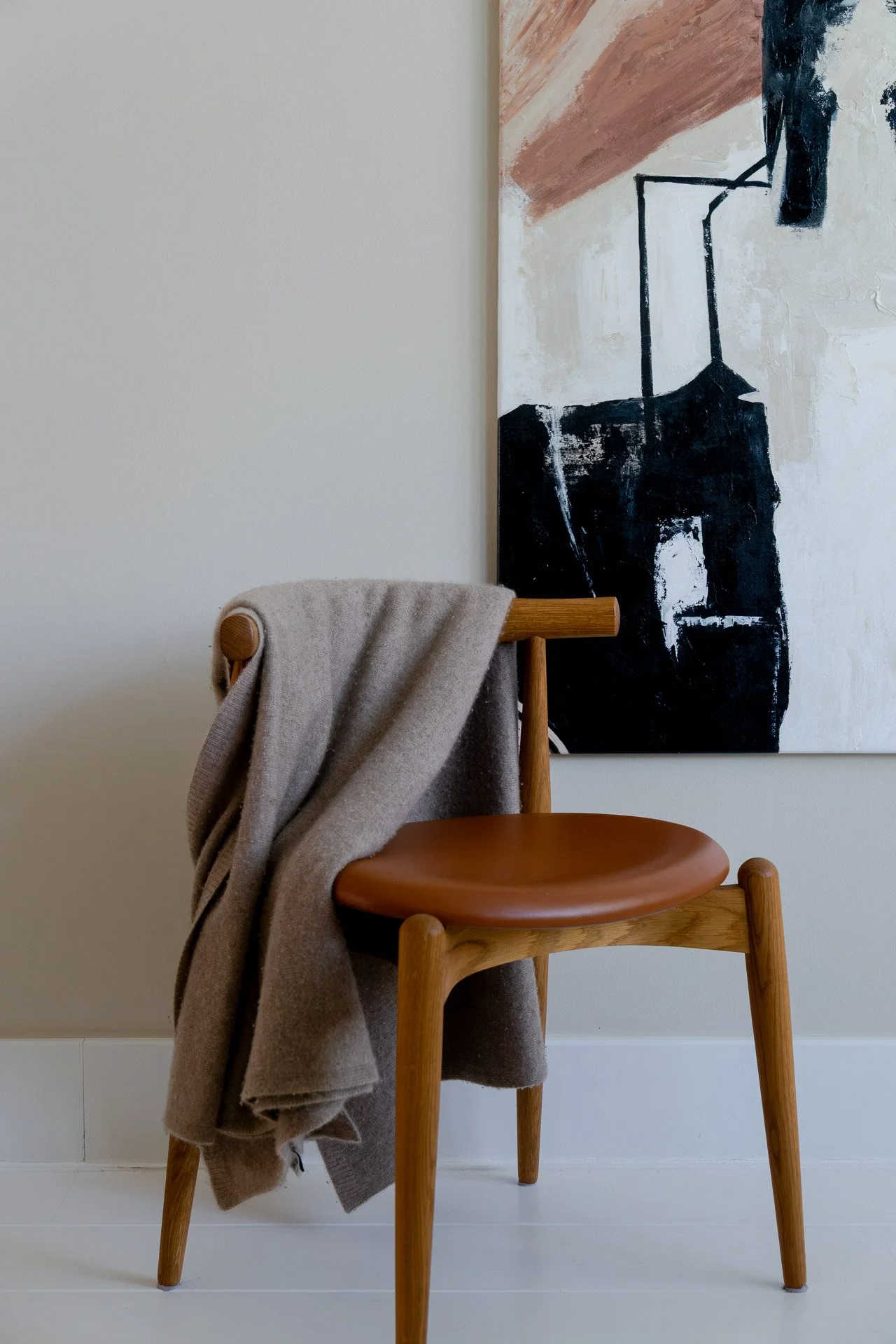 A wooden chair with a light brown cushioned seat, beige fabric draped over the backrest, positioned in front of a large abstract painting with black, white, and brown brushstrokes on a white wall.