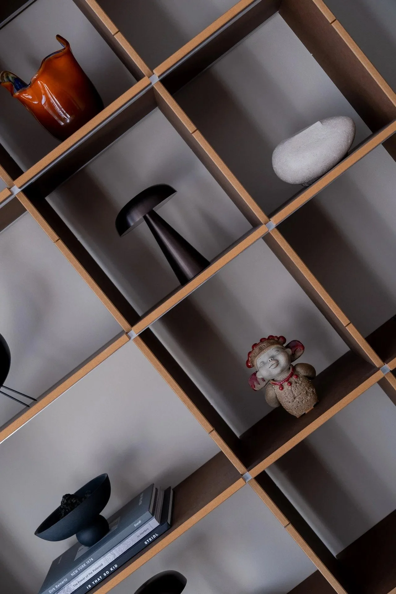 A black shelving unit with various decorative items. On the top left, a brown ceramic vase; on the middle right, a smooth flat stone; on the middle bottom, a small ceramic figurine of a smiling girl with a red bow; on the bottom right, a stack of books with a black vase and a speaker.
