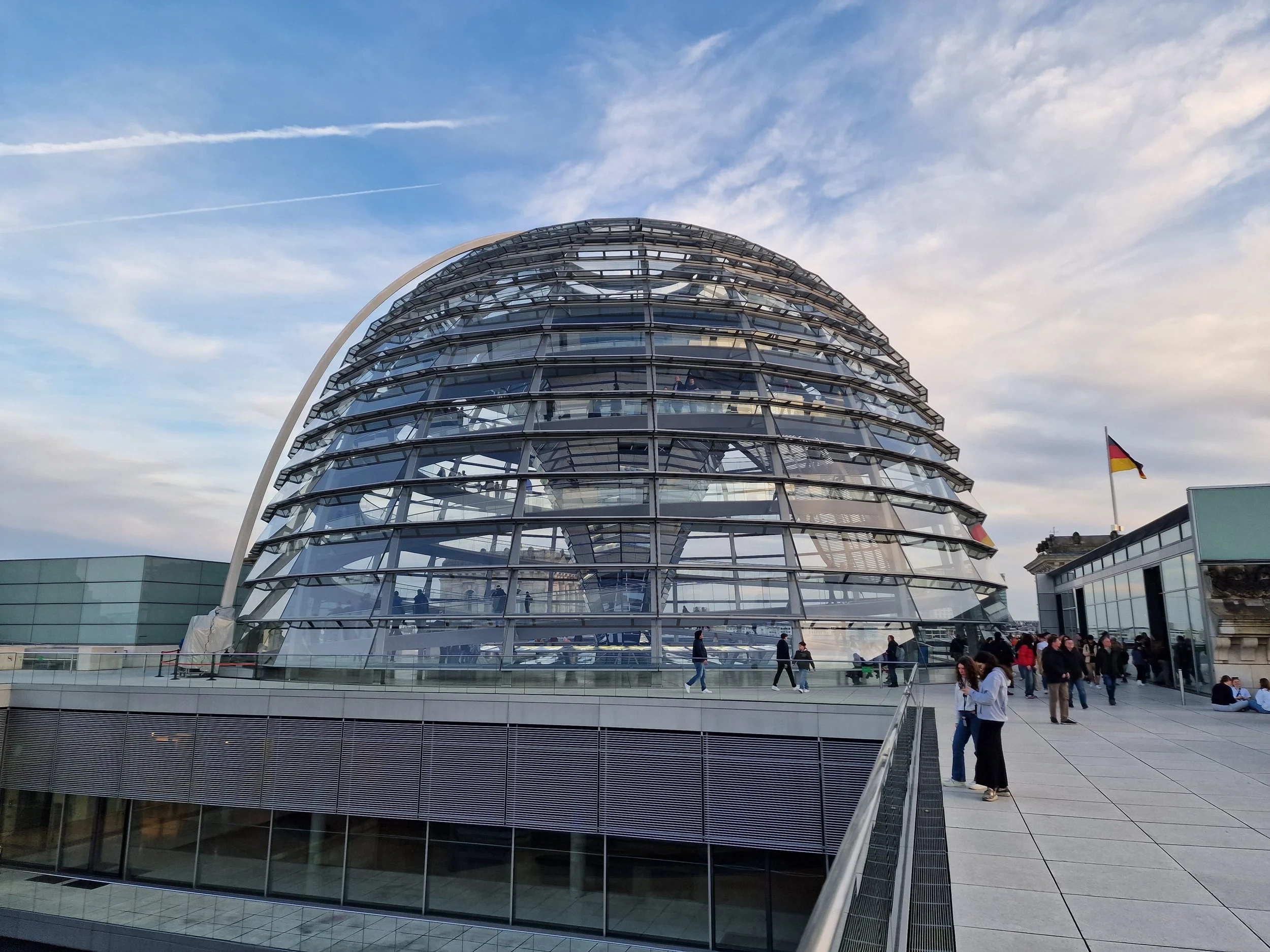 Das Bild zeigt ein modernes, spiralförmiges Glasgebäude mit mehreren Stockwerken, unter einem blauen Himmel mit Wolken. Es sind Menschen auf einer Plattform im Vordergrund zu sehen, einige sitzen und andere gehen.