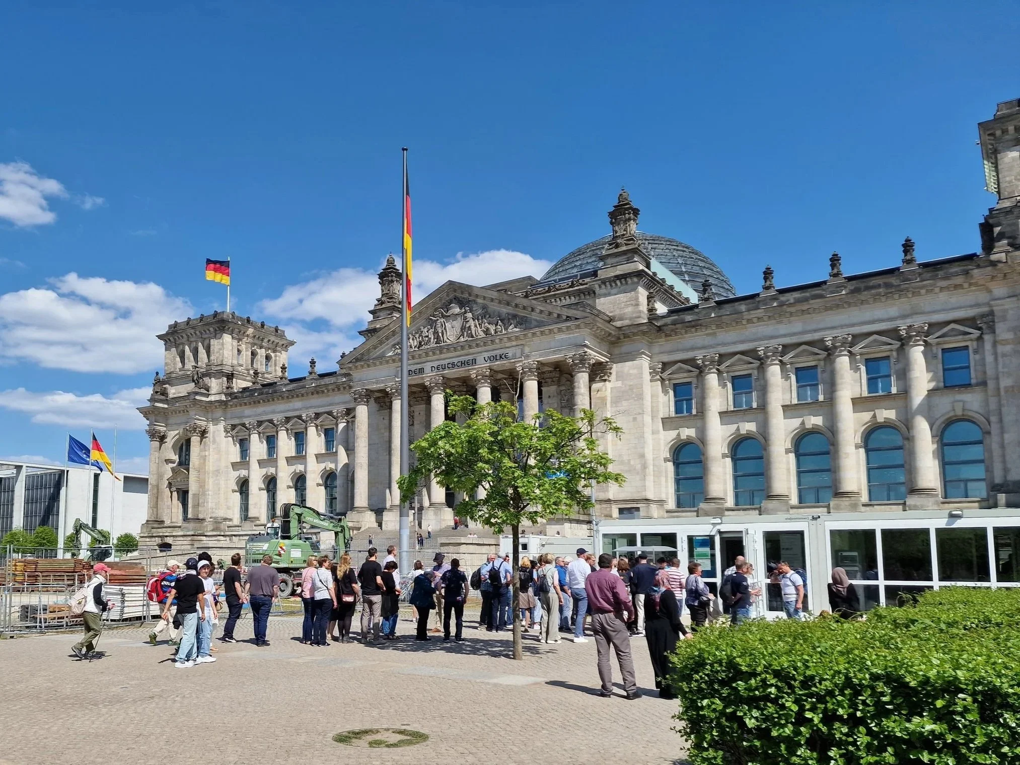 Das Bild zeigt eine große historische Regierungsgebäude mit einer Kuppel und mehreren deutschen Flaggen auf dem Dach. Vor dem Gebäude stehen viele Menschen, einige warten in einer Schlange, andere spazieren vorbei. Es gibt Bäume und eine Baustelle im Vordergrund sowie eine Linie von Bussen auf der rechten Seite.