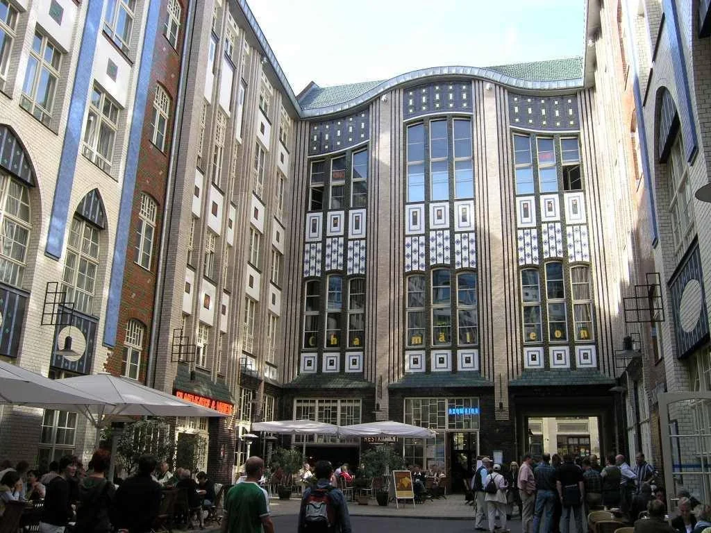 Aufnahme eines Innenhofs mit mehreren Leuten, die vor einem modernen Gebäude sitzen oder stehen, mit Tischen und Sonnenschirmen im Außenbereich.
