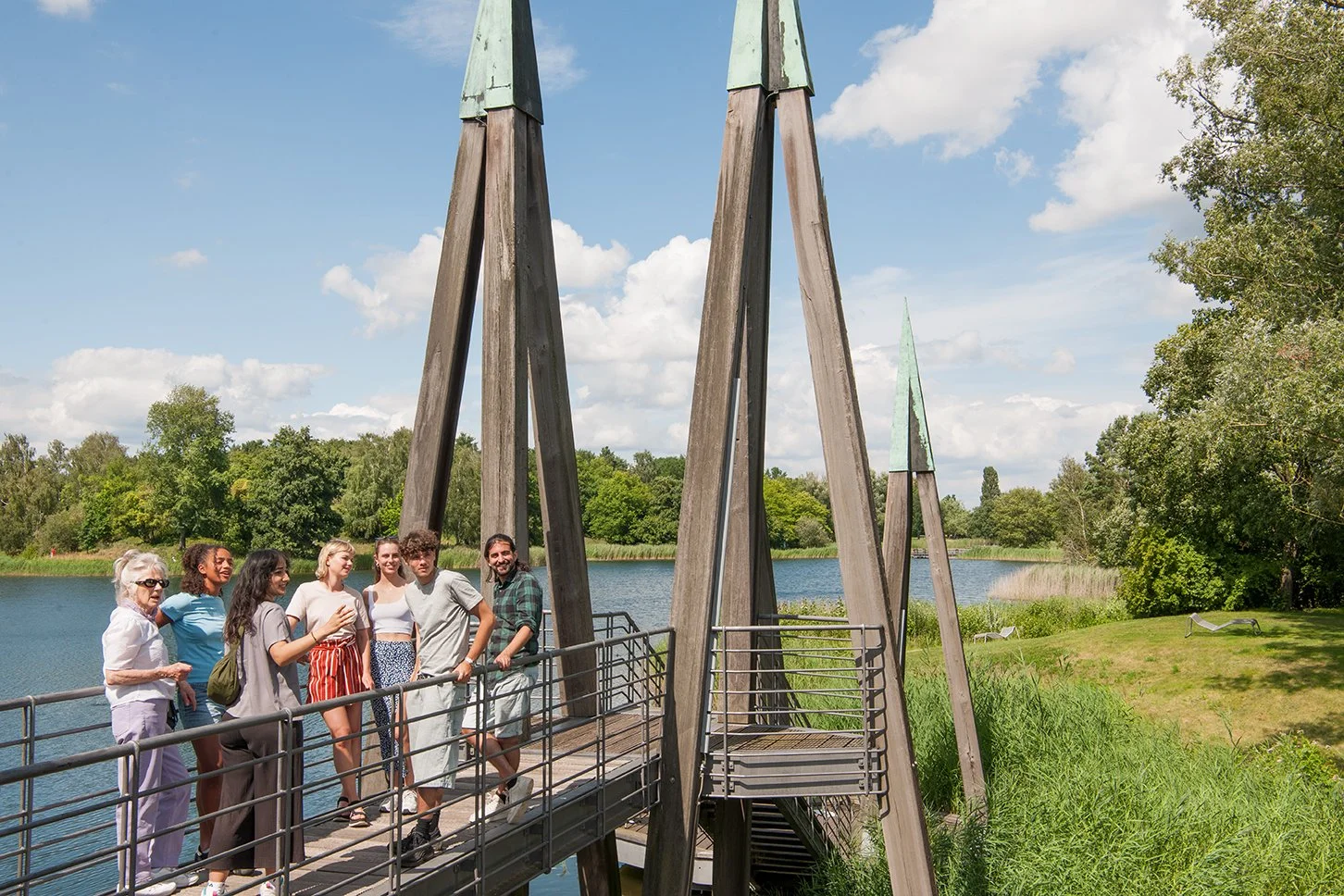 Eine Gruppe von acht Menschen steht auf einer Holzbrücke an einem See, umgeben von grünen Bäumen und natürlichen Strukturen, bei sonnigem Himmel.
