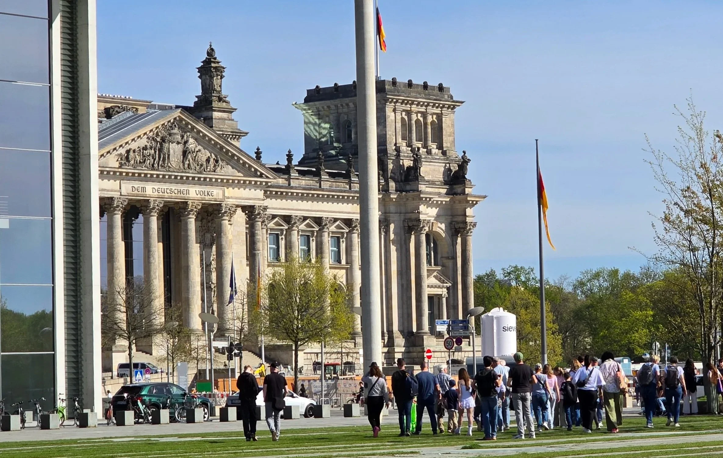 Das Bild zeigt den Reichstag in Berlin, Deutschland, mit Menschen, die vor dem Gebäude spazieren oder warten. Es gibt Flaggen, Bäume und eine Mischung aus moderner und historischer Architektur.
